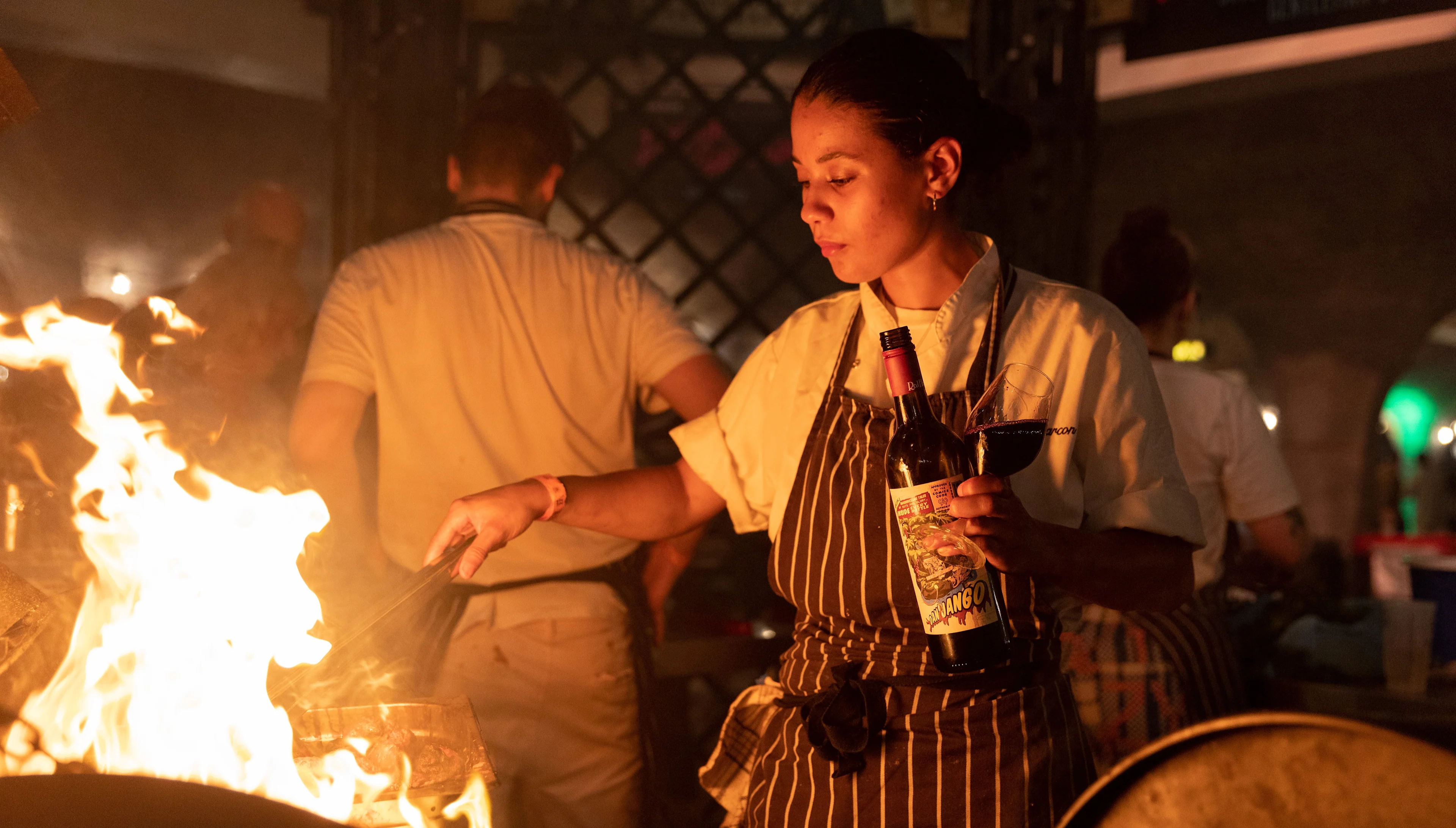 Chef cooking over an open flame while holding a bottle of wine at a nighttime event.
