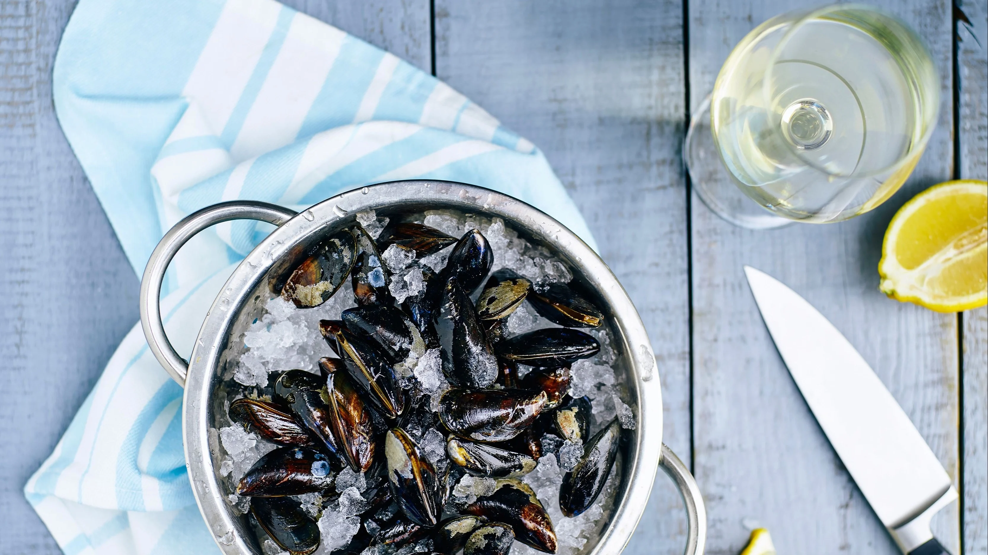 Bowl of fresh mussels on ice served with lemon wedges and a glass of white wine.