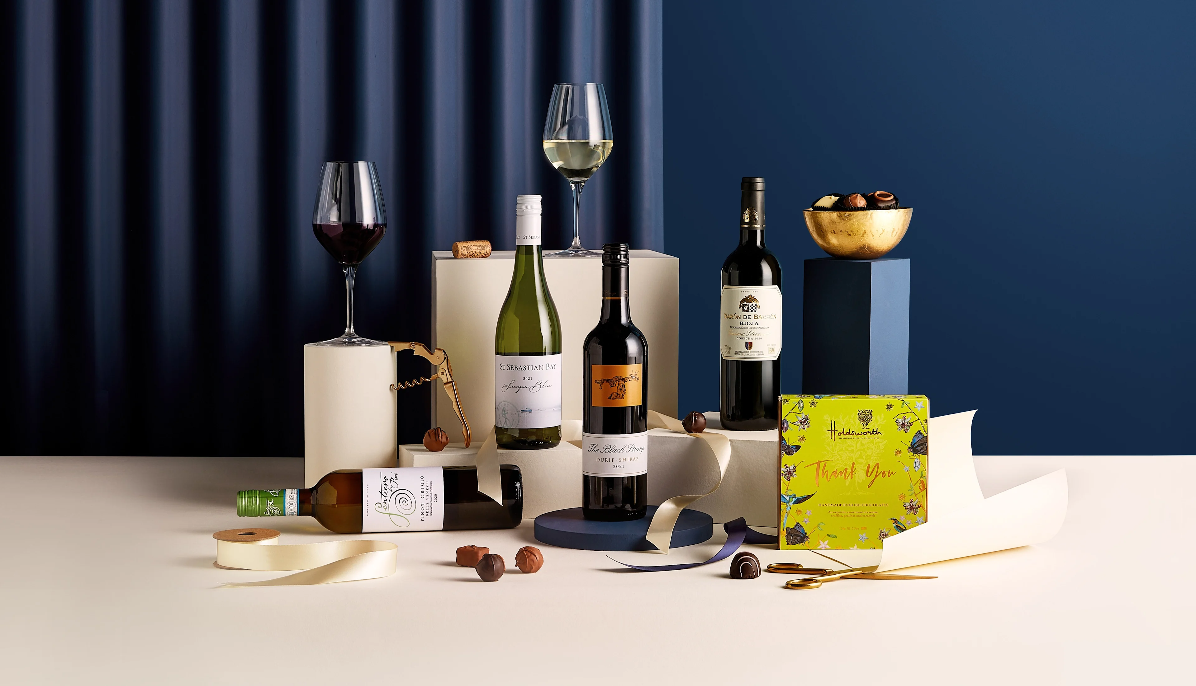 Selection of wine bottles, filled wine glasses, chocolates and gift wrap arranged on display blocks against a dark blue backdrop.