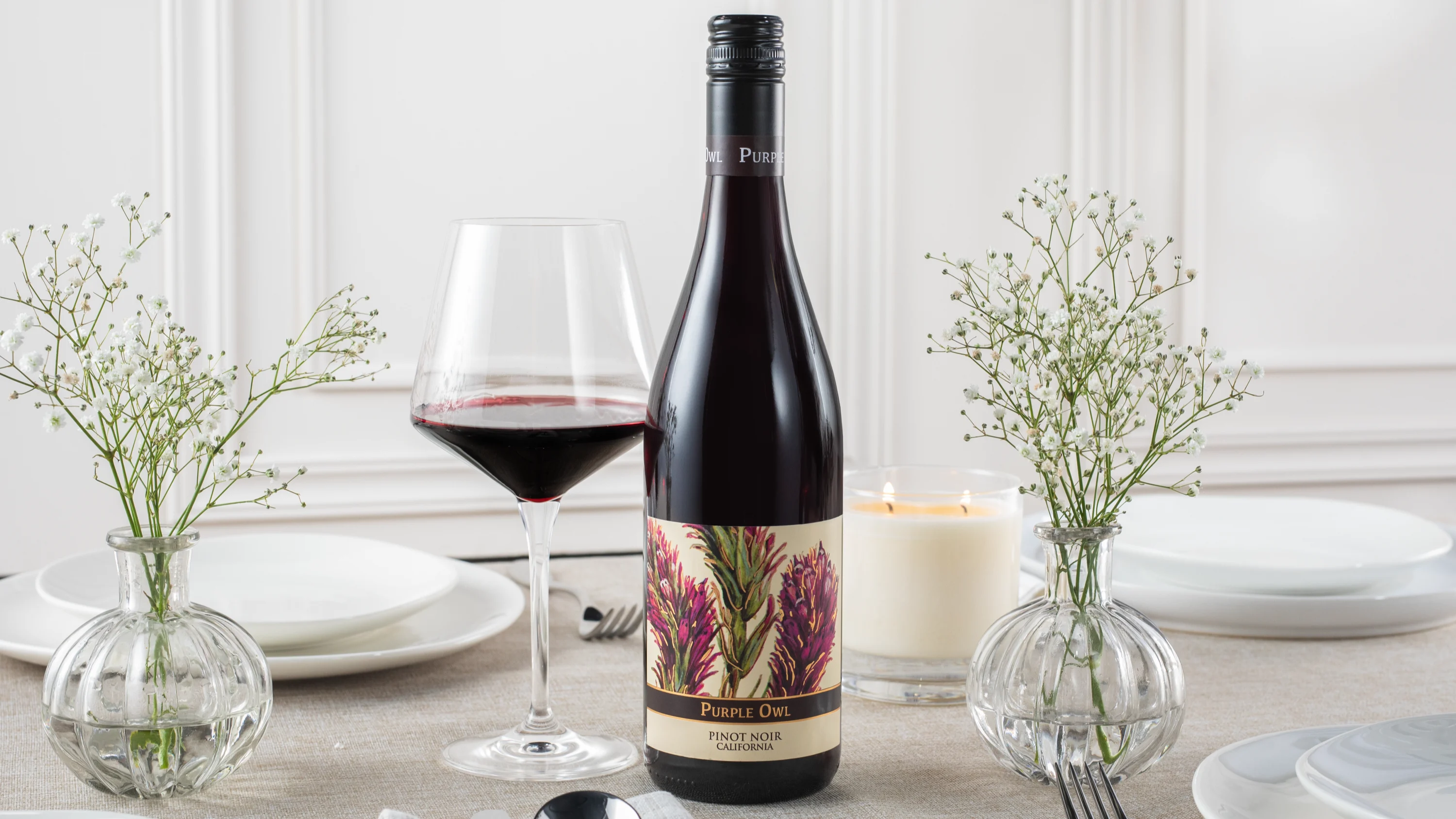Bottle and glass of Pinot Noir displayed on a table set with plates, candles and small floral arrangements.