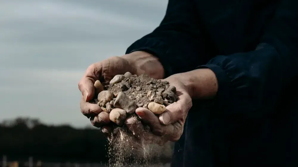 A close-up shot of a pair of hands holding soil and rocks, with some soil passing through their fingers.