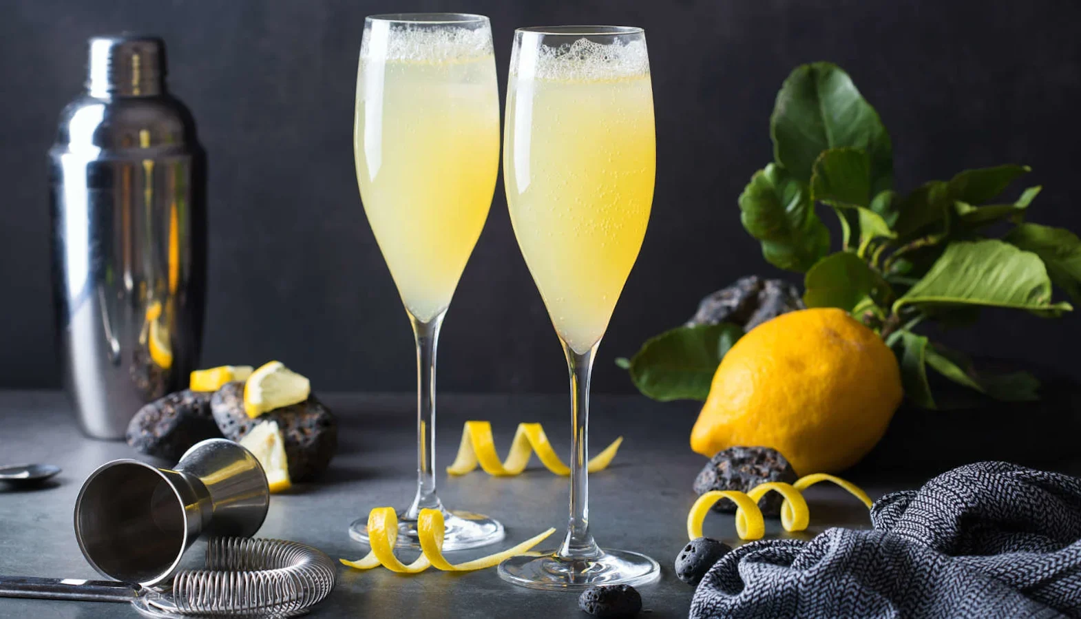 Two champagne flutes with pale yellow cocktail, lemon twists, and a shaker in the background.