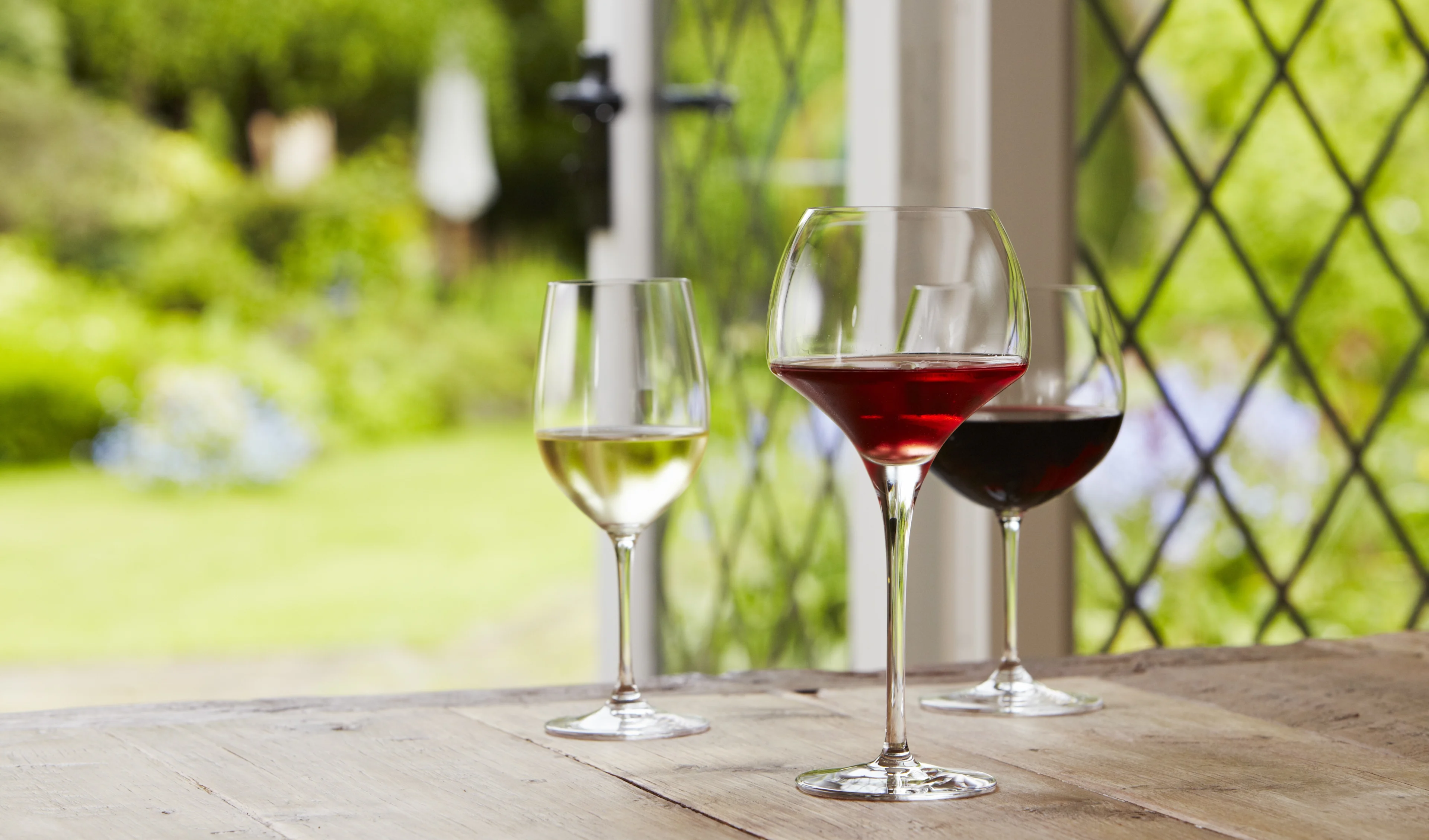 Three glasses of red and white wine on a wooden table beside open doors looking out onto a garden.