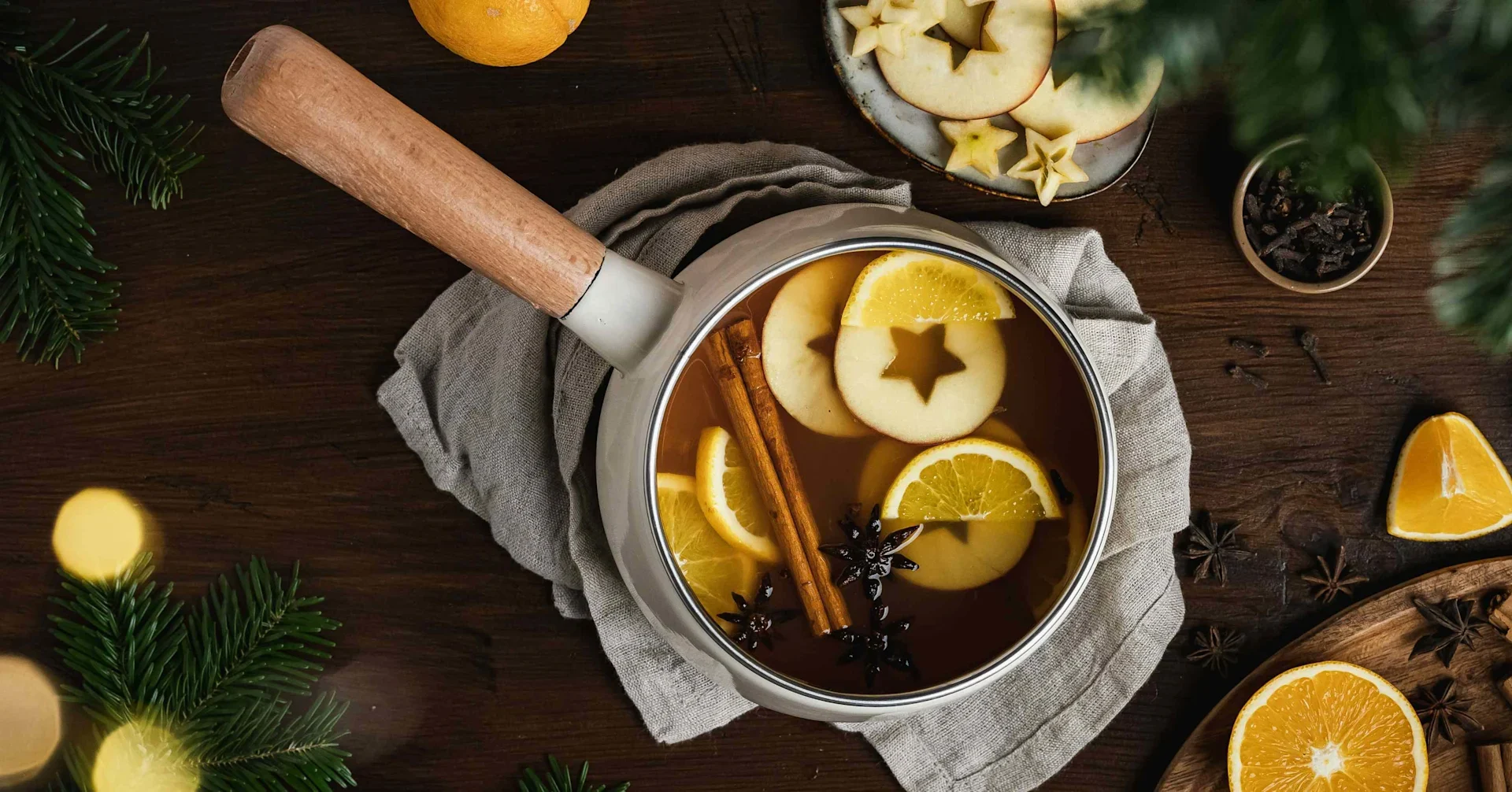 Saucepan of mulled cider with orange slices, star-shaped apple slices, cinnamon sticks, and star anise on a wooden table.