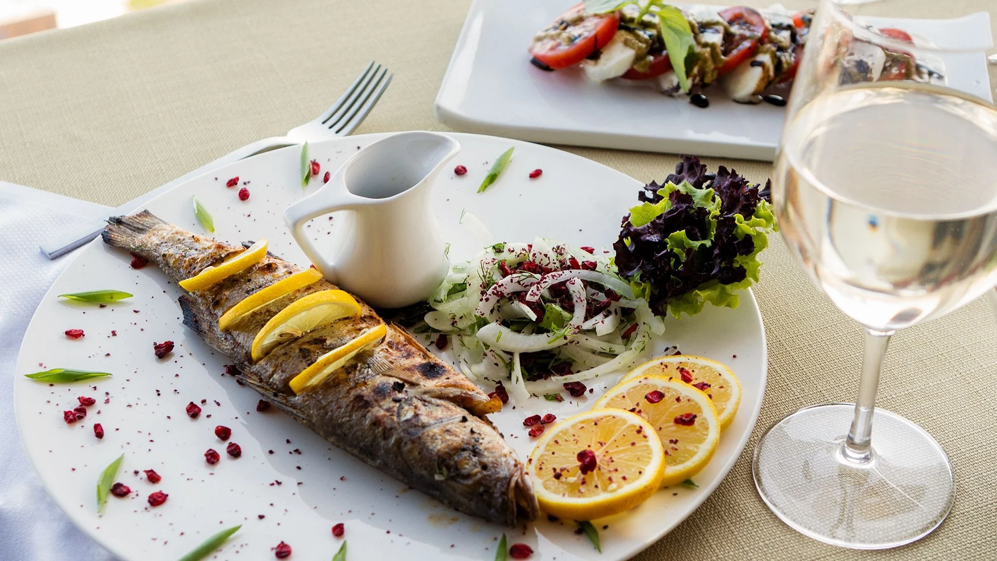 Grilled whole fish with lemon slices served alongside salad and a glass of white wine.
