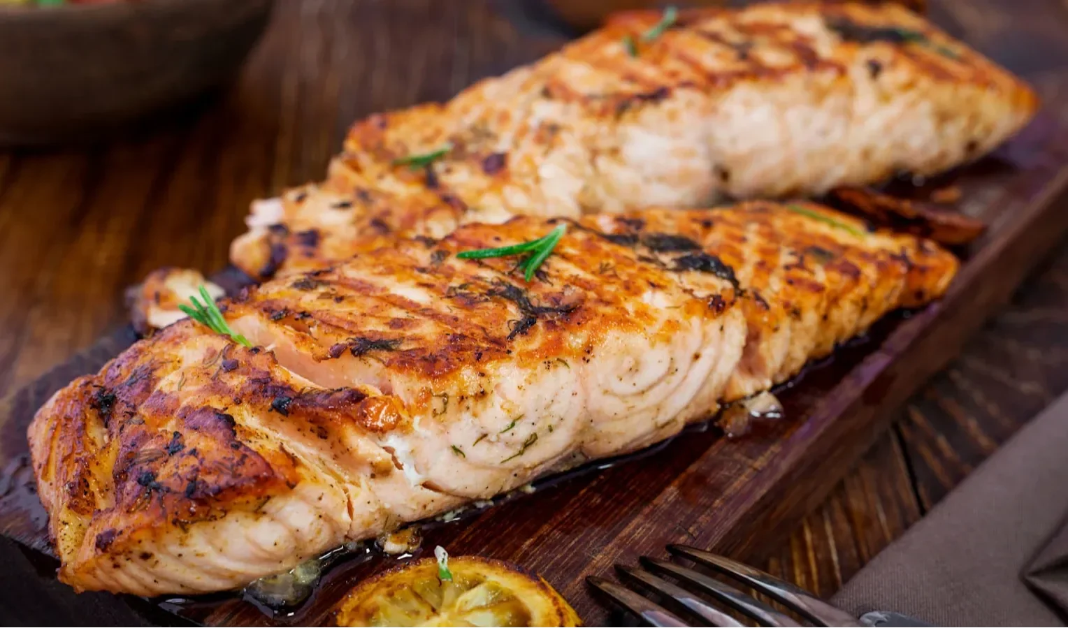 Grilled salmon fillets on a wooden board with herbs and charred lemon slices.
