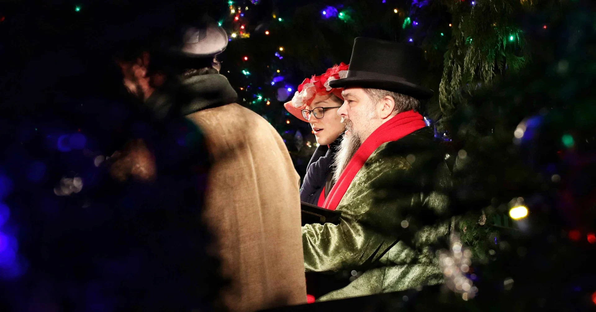 People in festive clothing seated among Christmas lights and greenery during a holiday event.
