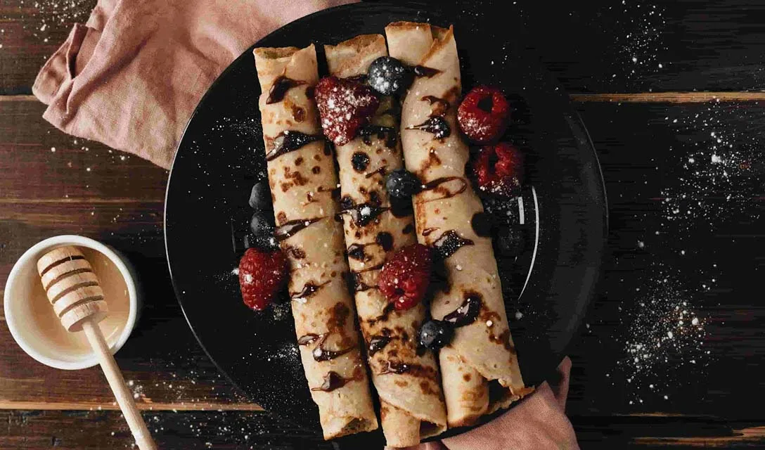 Rolled crepes topped with raspberries, blueberries, and chocolate drizzle on a black plate.