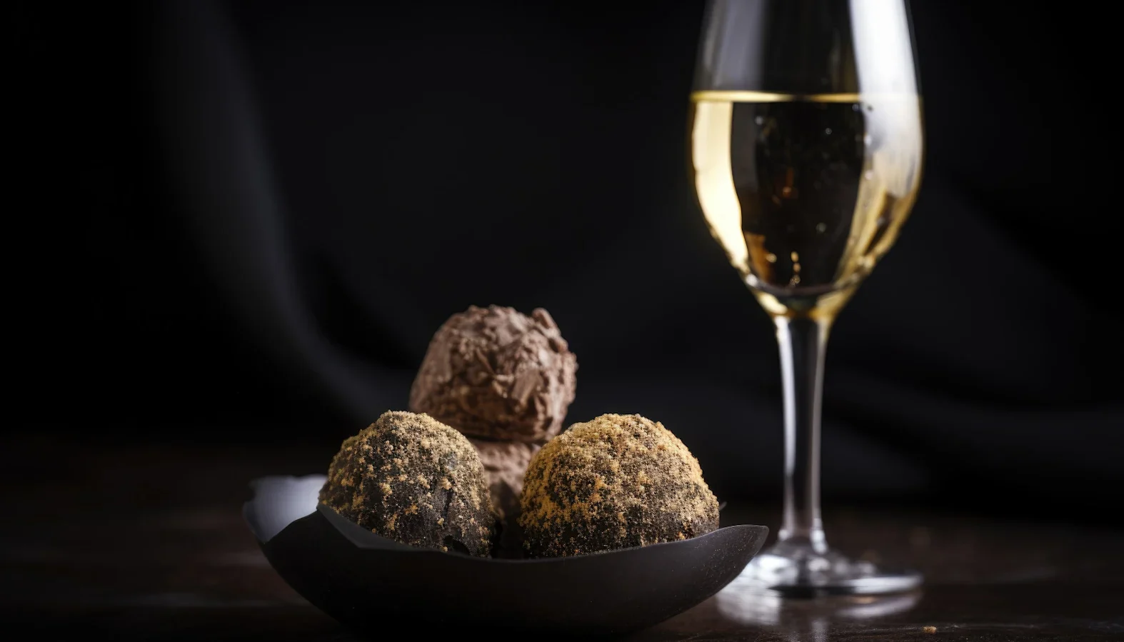 Plate of chocolate truffles beside a glass of sparkling wine against a dark background.