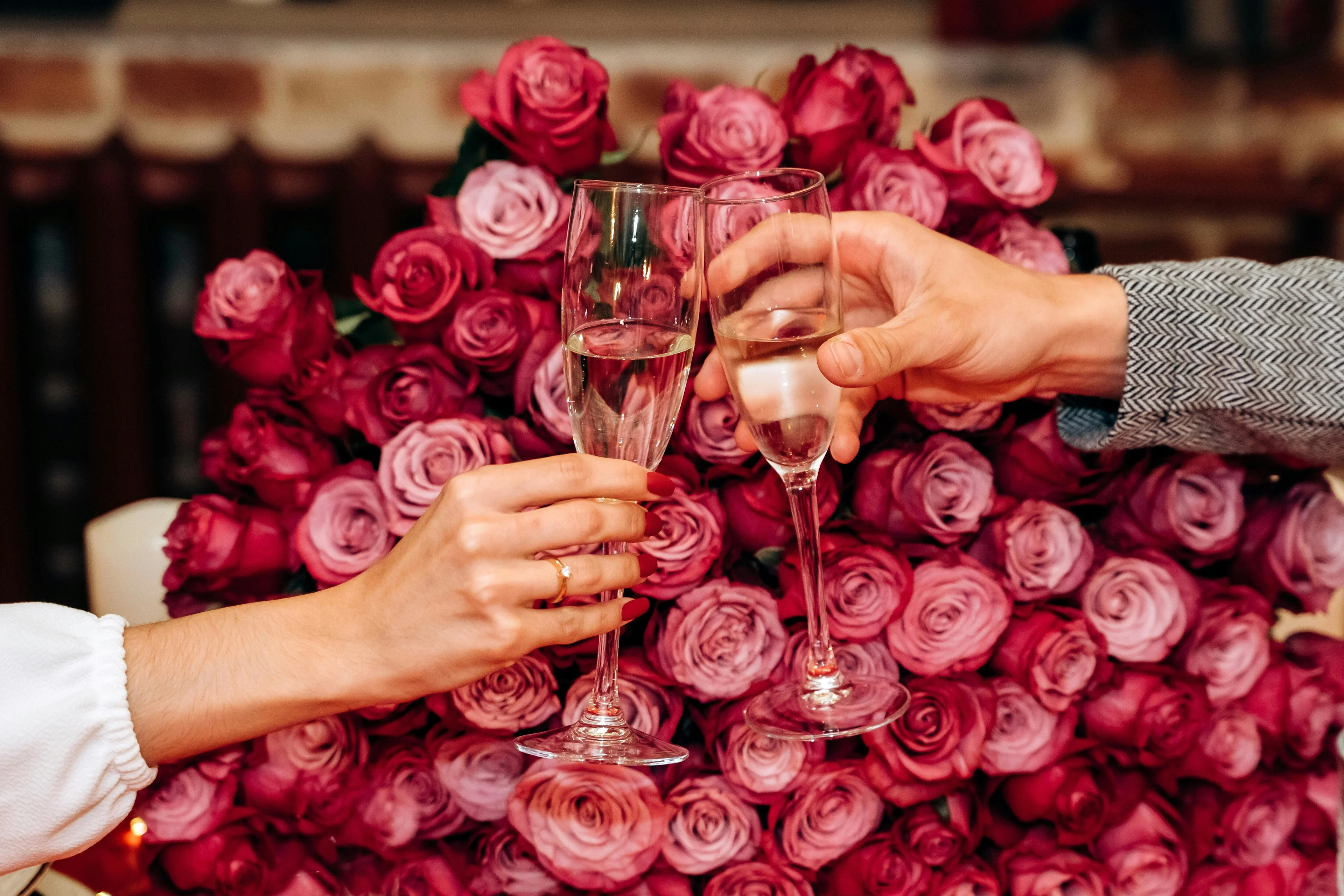 Two hands clinking champagne flutes filled with prink champagne in front of pink roses.