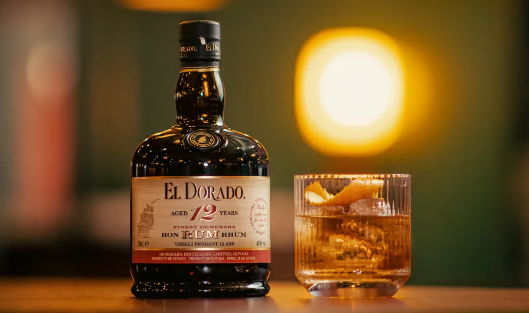 Bottle of El Dorado 12-Year Rum next to a glass of rum cocktail with citrus peel on a dark wooden surface under warm lighting.