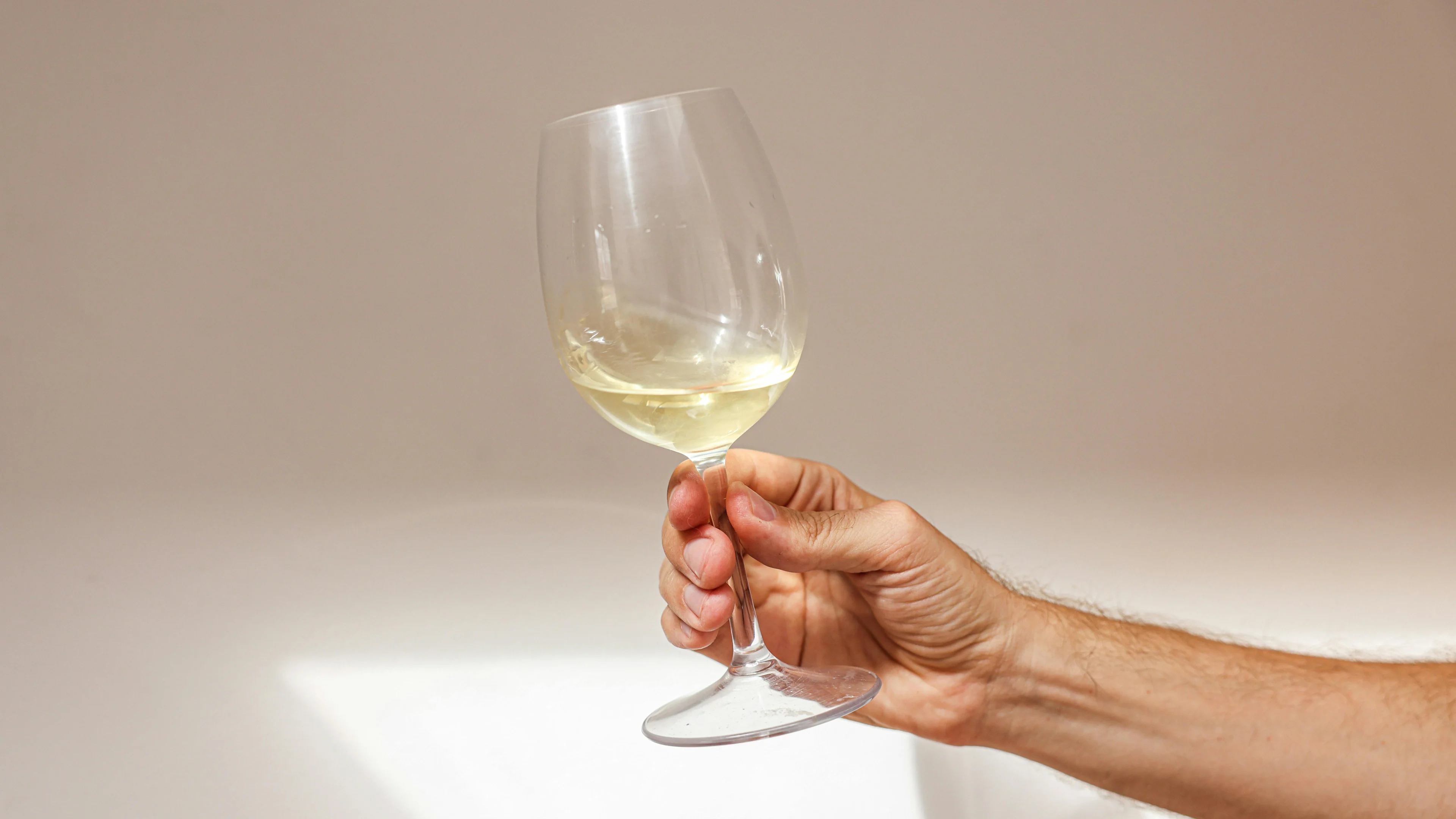 Hand holding a glass of pale white wine against a plain light-coloured background.