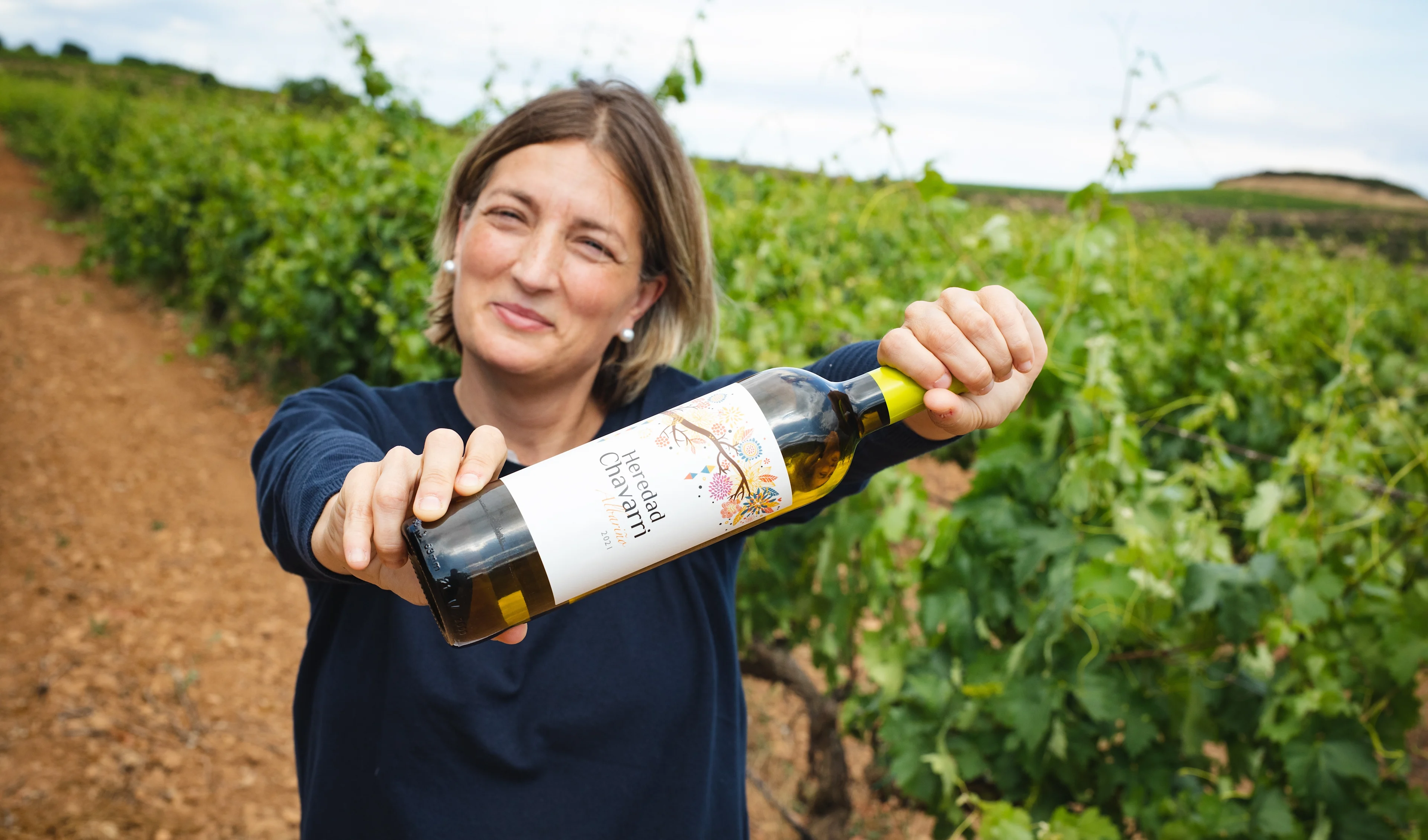 Person holding a bottle of Albariño wine while standing between vineyard rows.