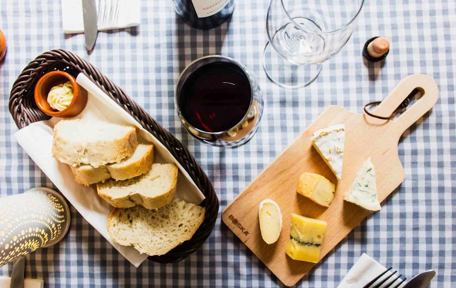 A small wooden cheeseboard with assorted cheeses next to a basket of sliced bread, a glass of red wine, and an empty wine glass on a checkered tablecloth.