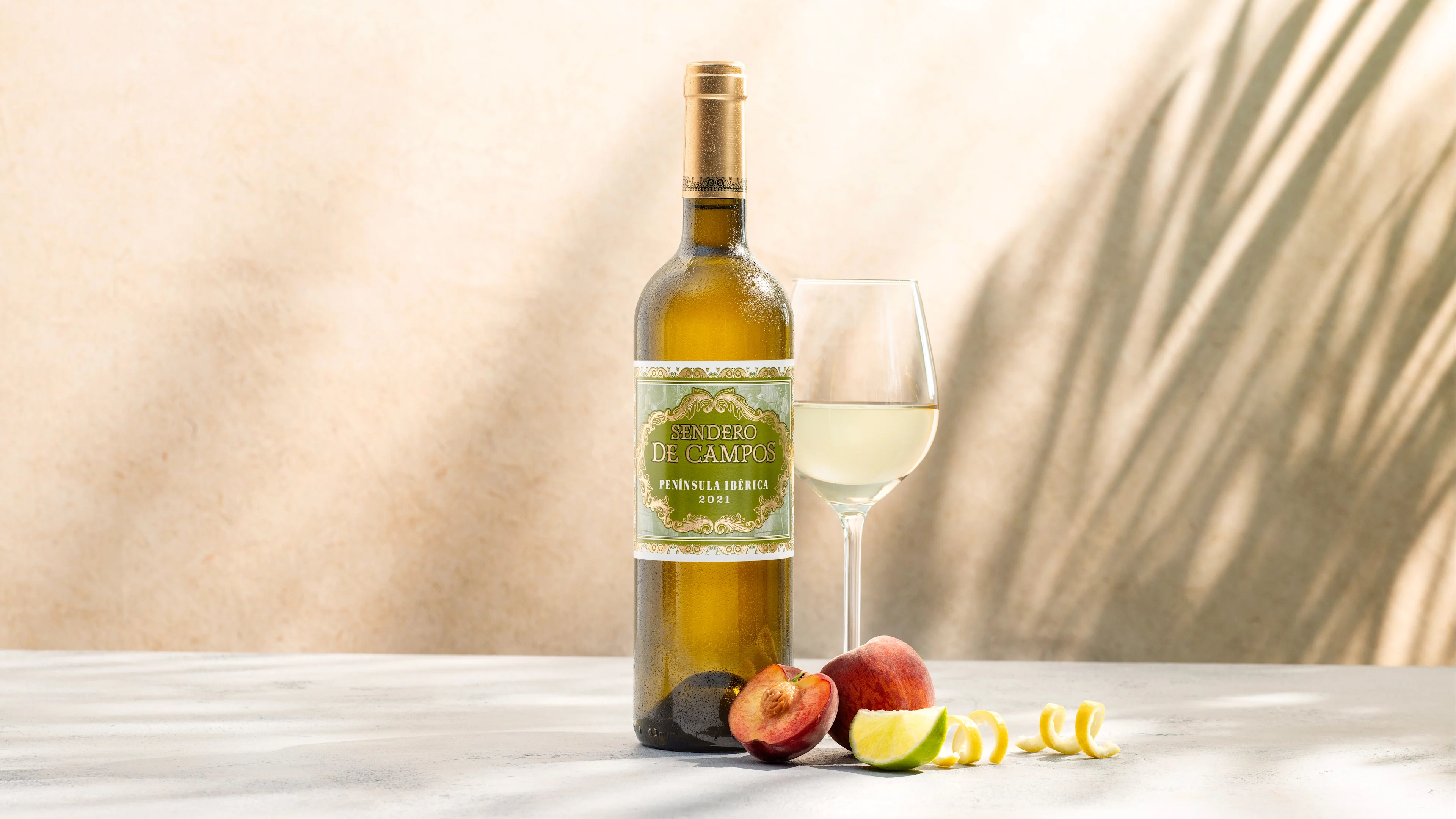 Bottle and glass of white wine beside sliced peach and citrus fruit against a softly lit backdrop.