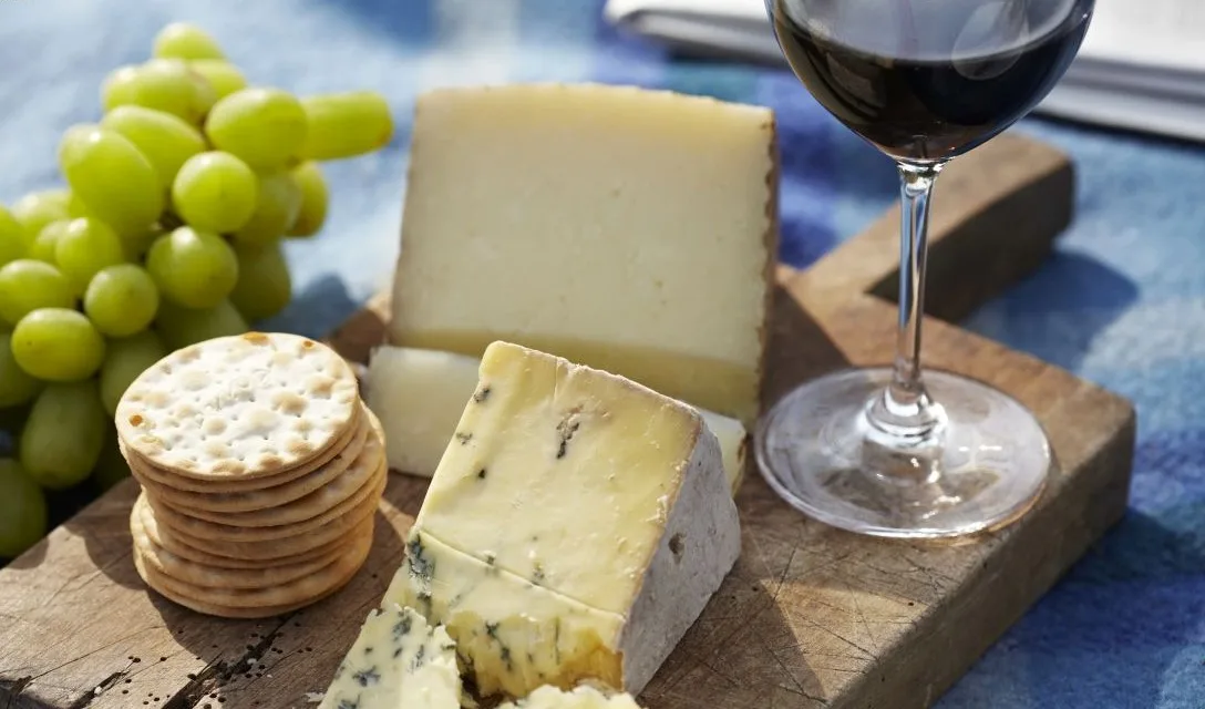 A wooden board with assorted cheeses, cured meats, and seeded crackers on an outdoor table, with several glasses of red and white wine and a person holding a glass of red wine.