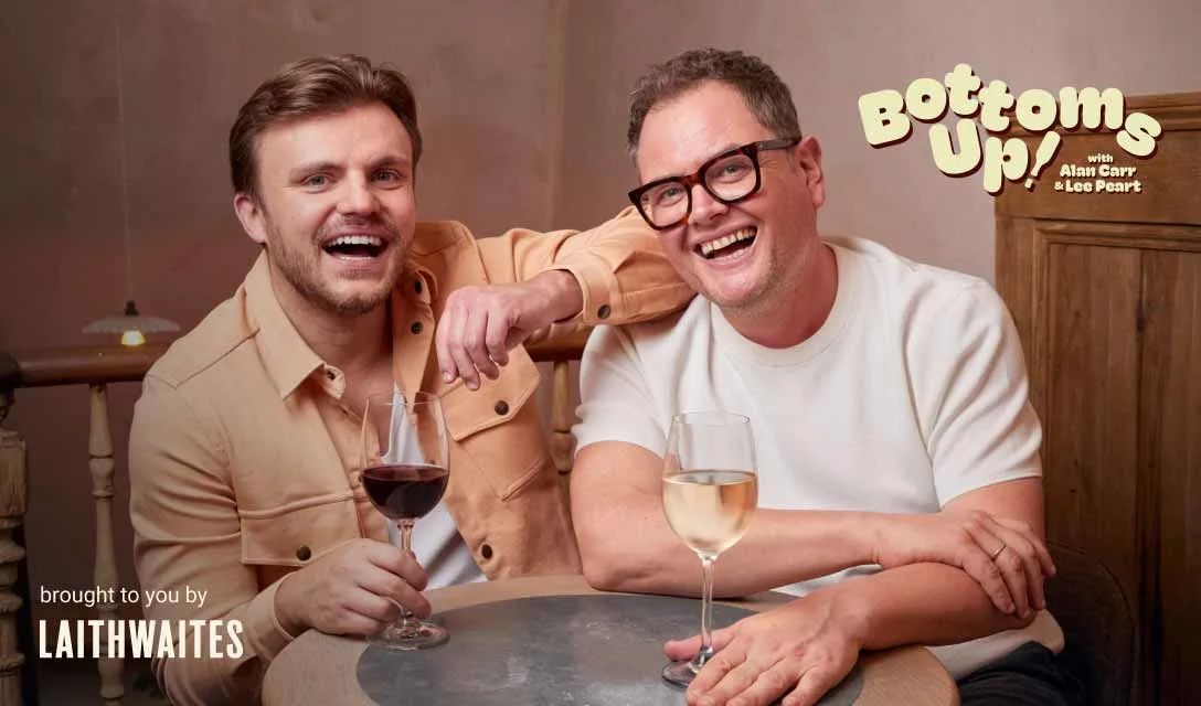 Alan Carr and Lee Peart sitting at a round table with glasses of wine, next to a sign that reads “Bottoms Up! with Alan Carr & Lee Peart,” sponsored by Laithwaites.