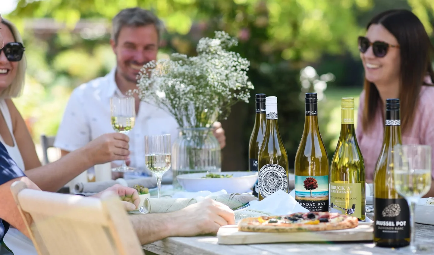 People gathered around an outdoor dining table with bottles and glasses of Sauvignon Blanc wine, food and flowers.
