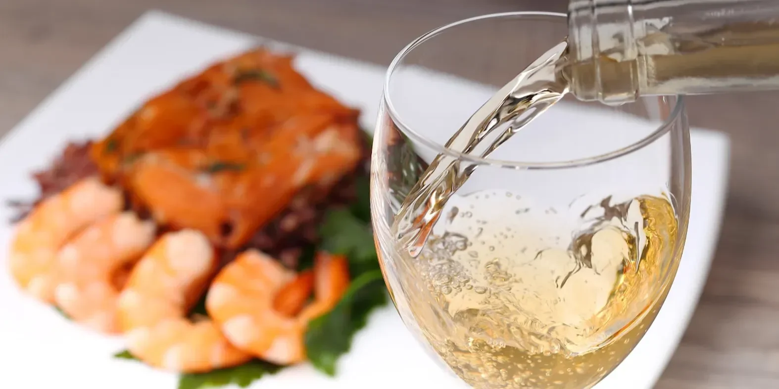 Glass of white wine being poured with a plate of salmon and prawns blurred in the background.