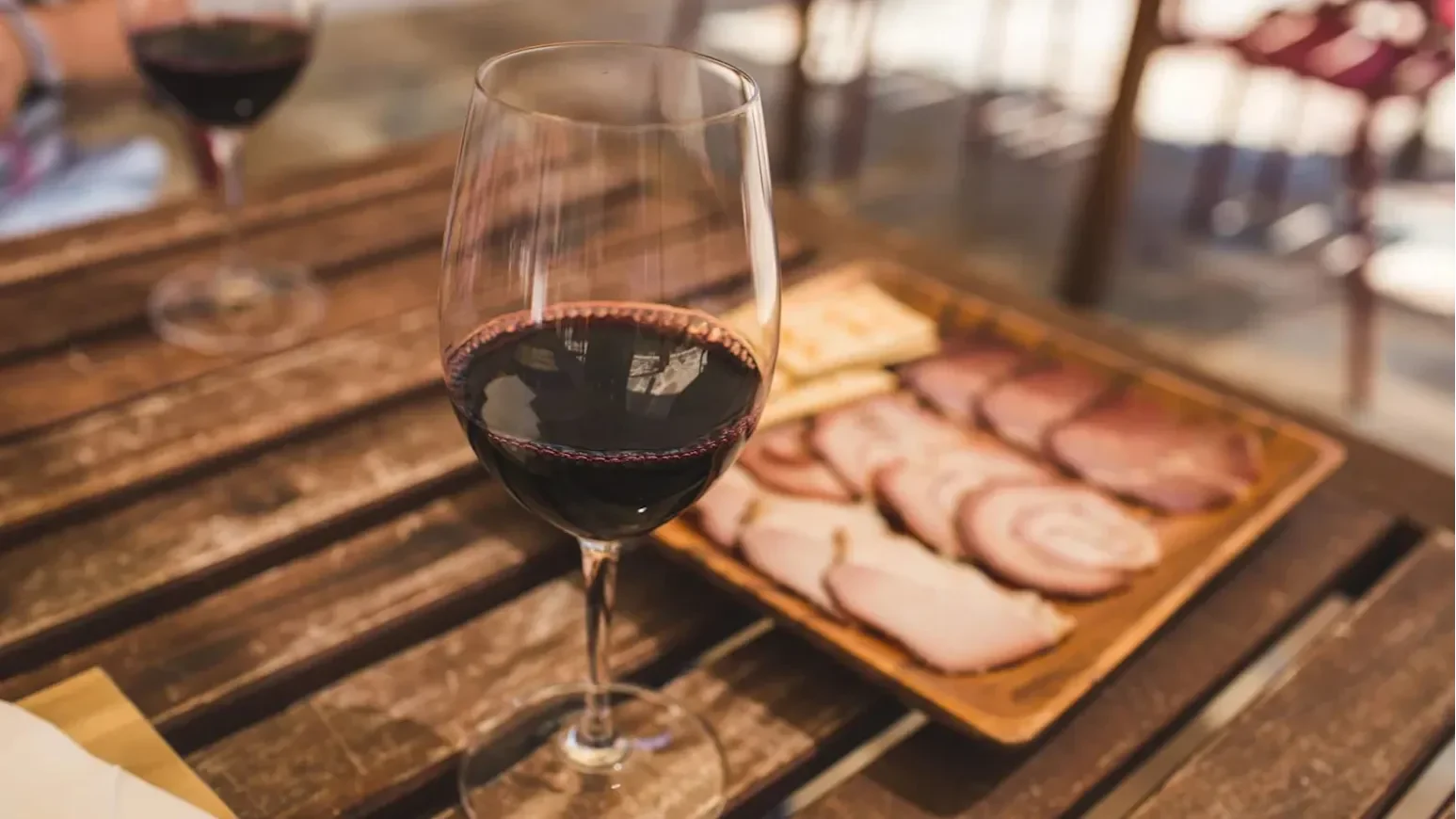 Glass of red wine on a wooden table with sliced cured meats on a serving board.