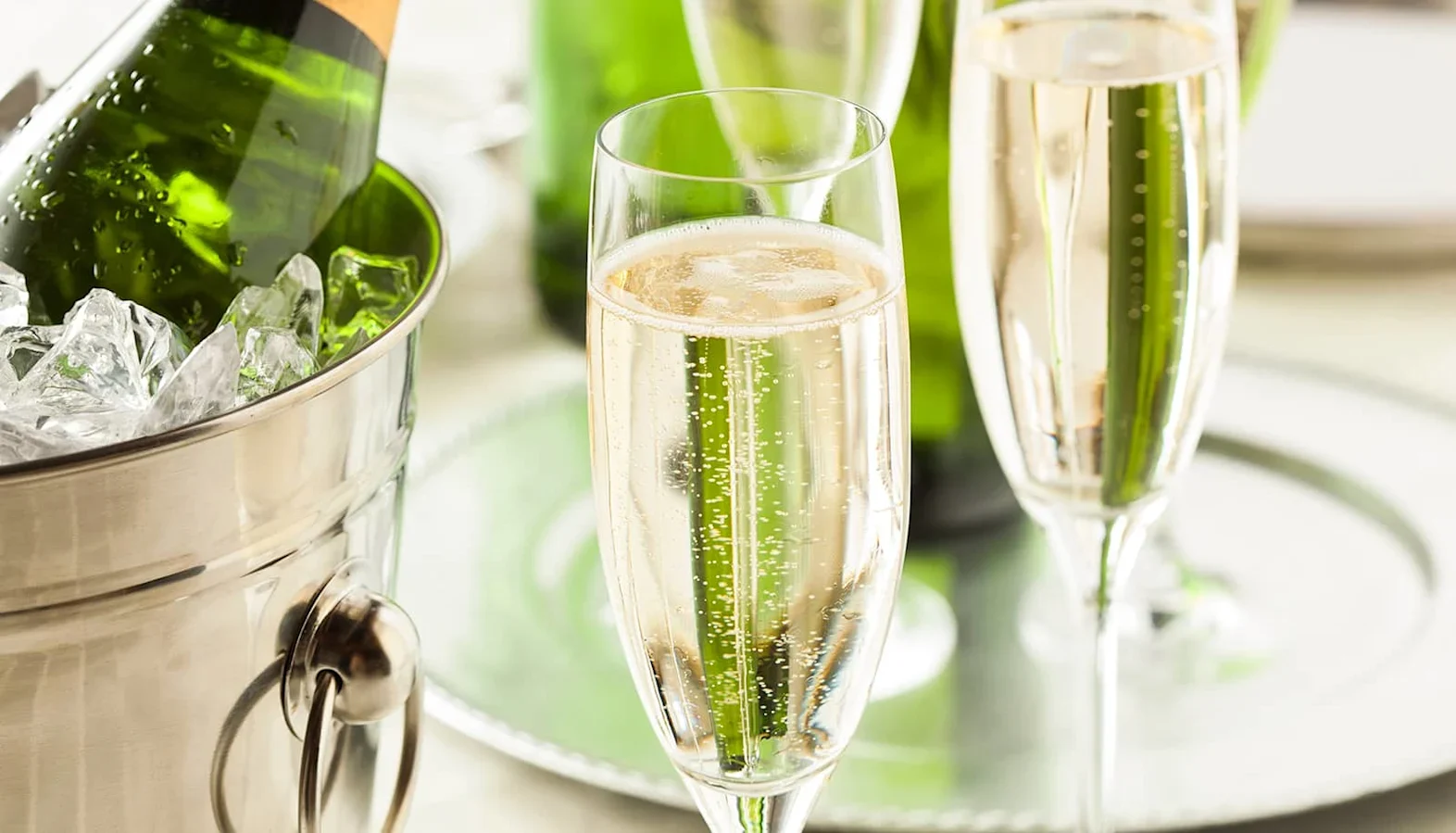 Champagne flutes filled with sparkling wine beside a metal ice bucket holding a green bottle on a reflective silver tray.