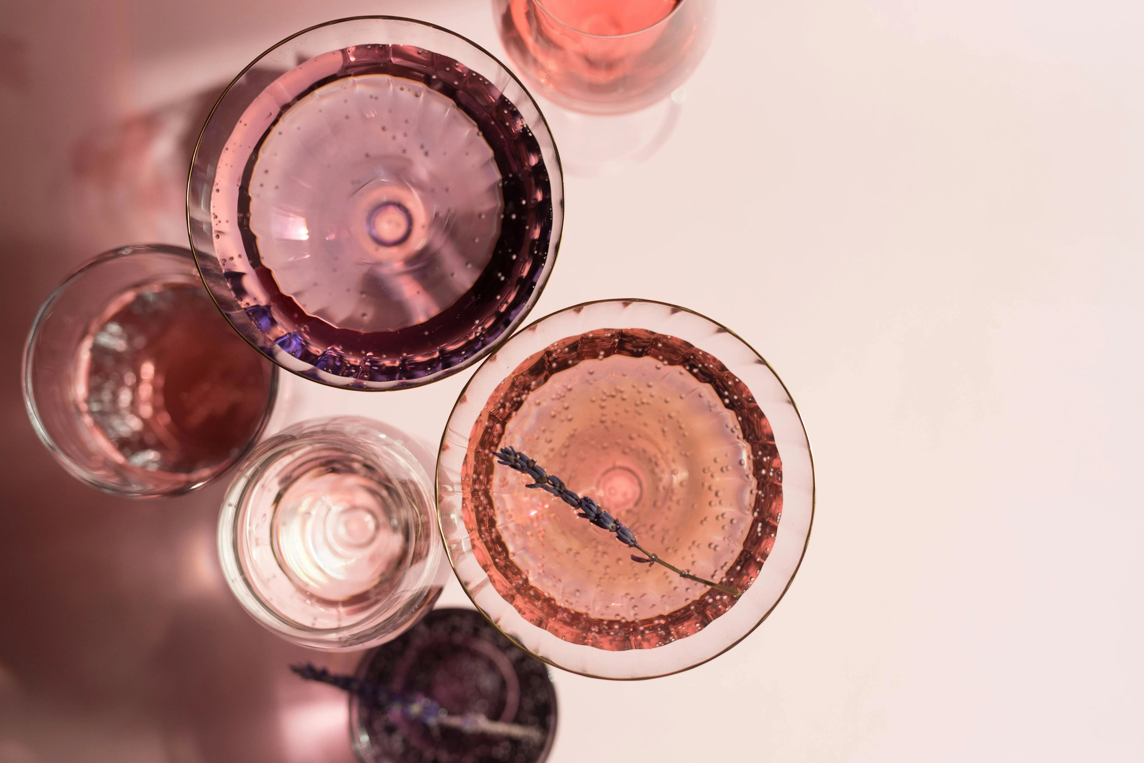 Overhead view of assorted wine glasses filled with pink drinks on a pale background, one has a sprig of lavender for garnish.