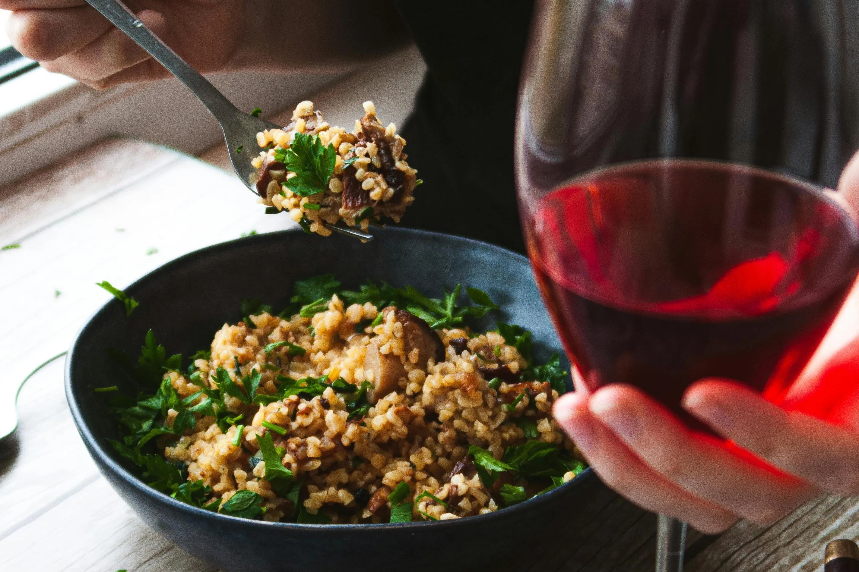 A person holding a glass of red wine while eating a bowl of cooked grains mixed with herbs and vegetables, using a fork, on a wooden table with a blue napkin.