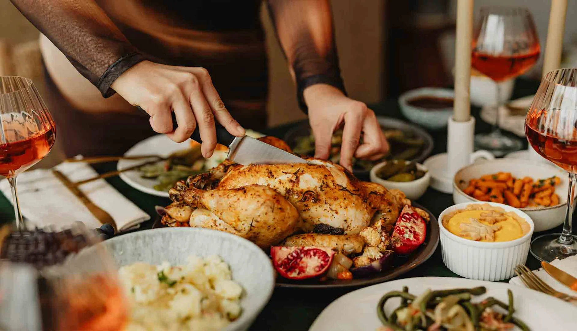 Hands carving a roast chicken on a table filled with side dishes and glasses of rosé wine.