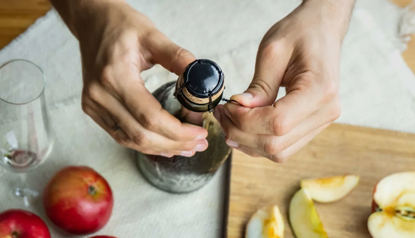 Hands opening the wire cage on a sparkling wine bottle, with apples, a glass, and sliced fruit arranged on a wooden board nearby.
