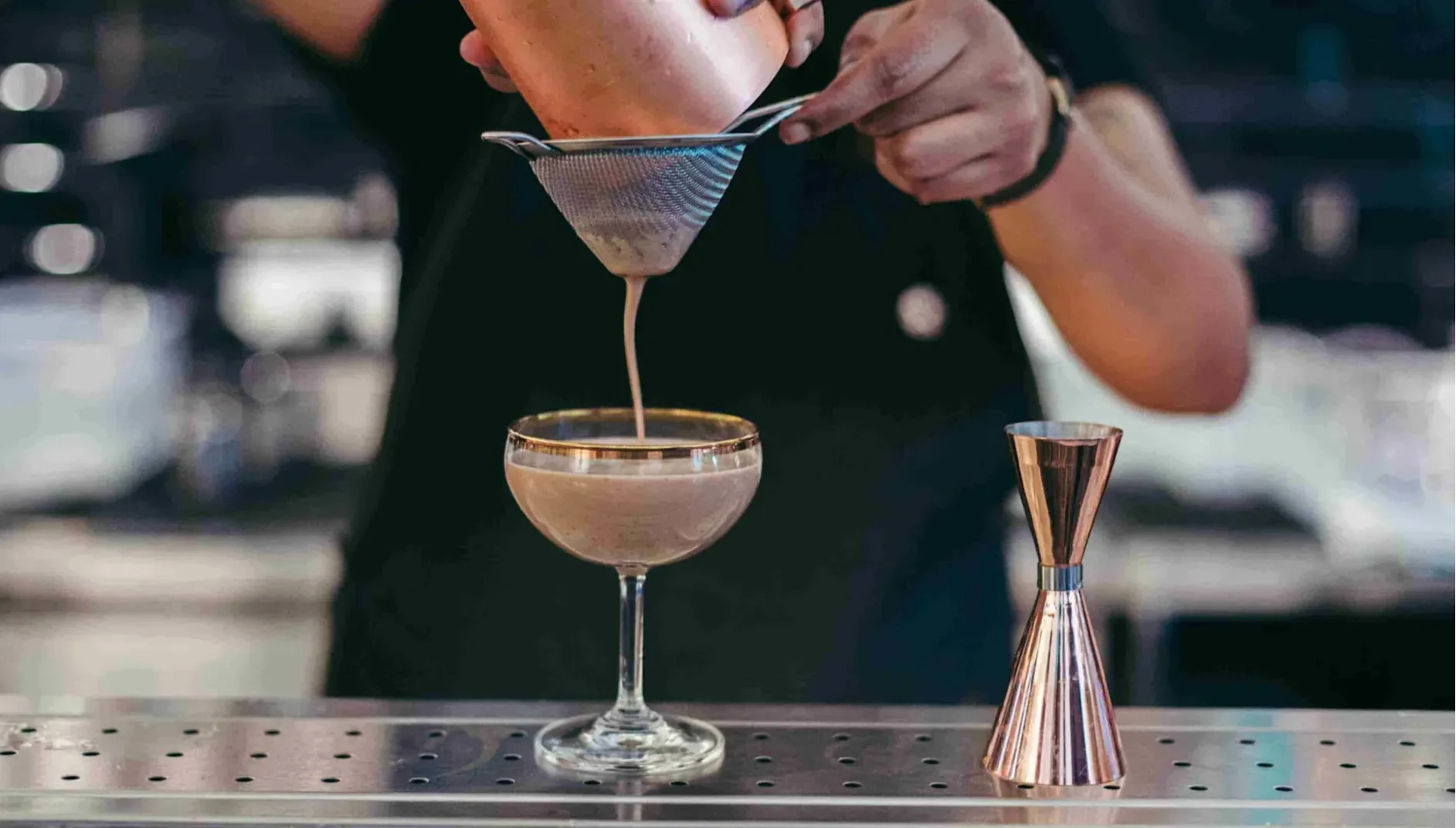 Creamy cocktail mixture being poured through a strainer into a coupe glass at a bar.