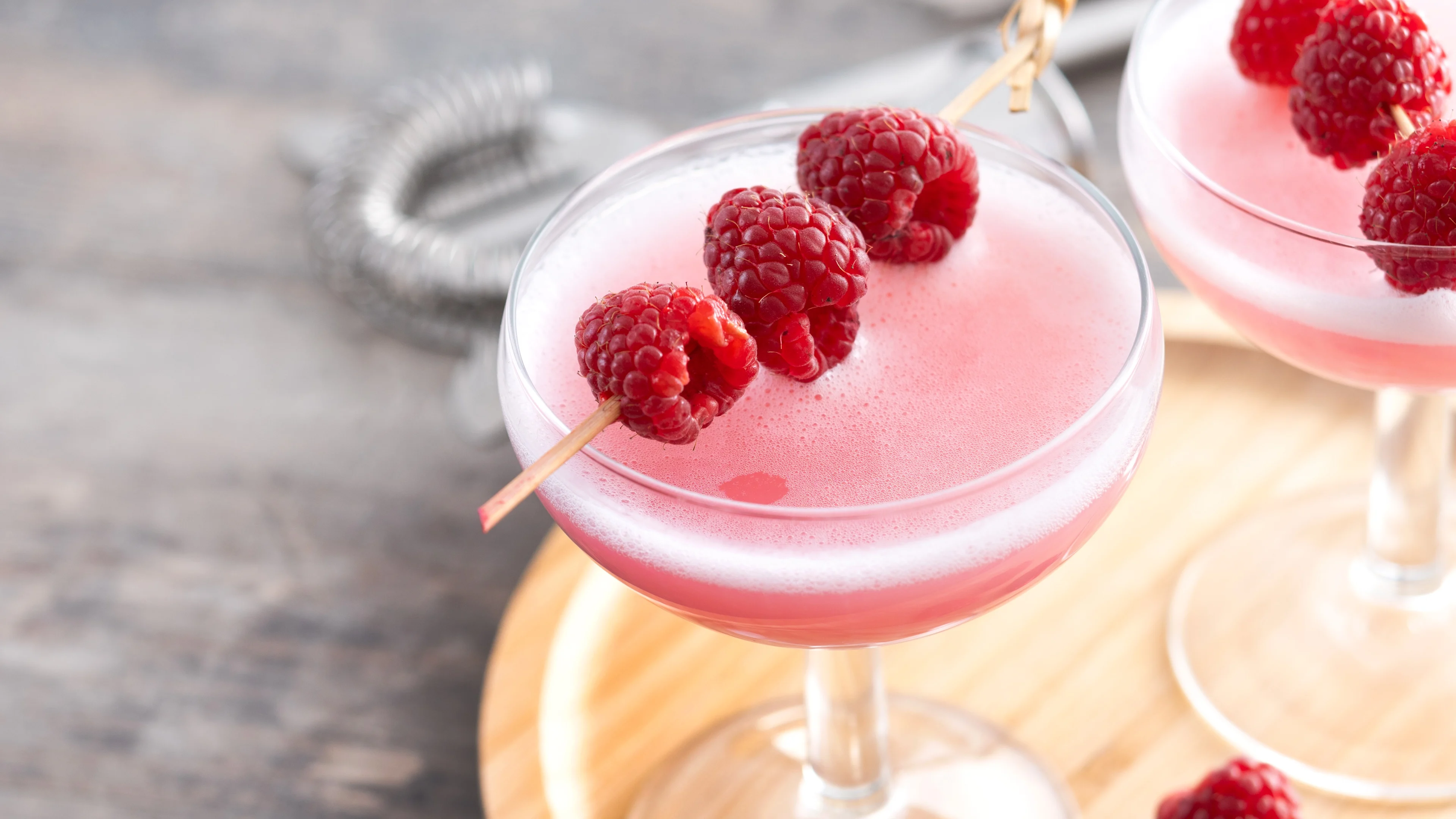A pink raspberry martini garnished with fresh raspberries on a stick on a wooden tray.