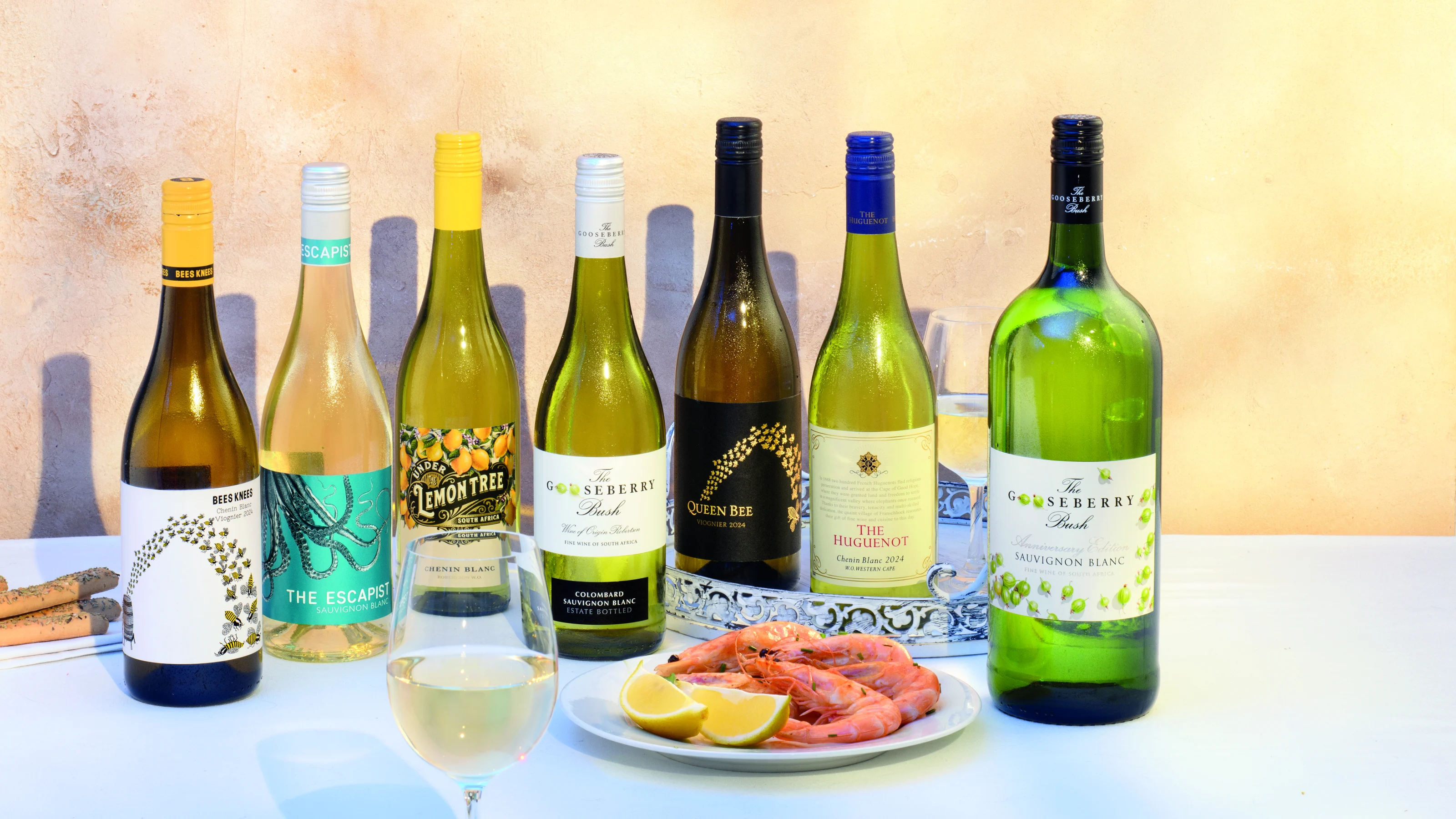 Selection of white wine bottles displayed with glasses of wine, lemon wedges and cooked prawns on a serving plate.