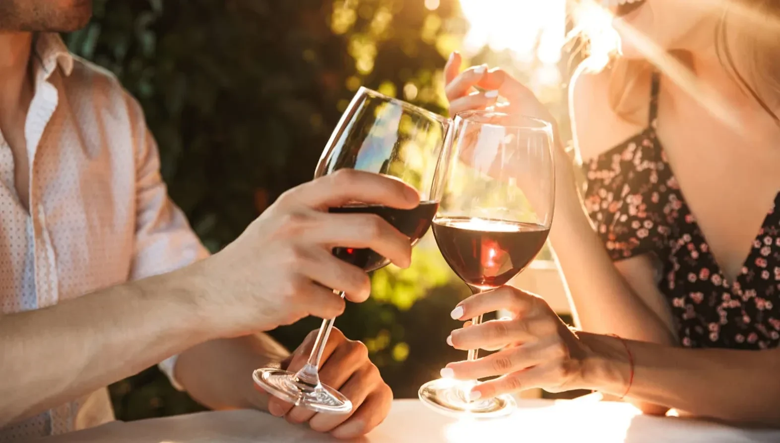 Two people clinking red wine glasses at a sunlit outdoor table.