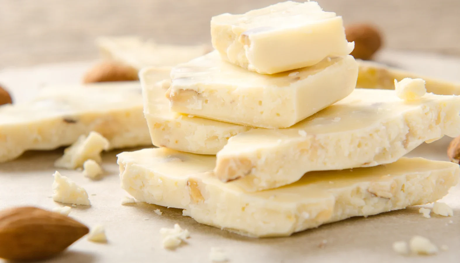 Stack of white chocolate pieces with nuts on a parchment surface, surrounded by scattered almonds.
