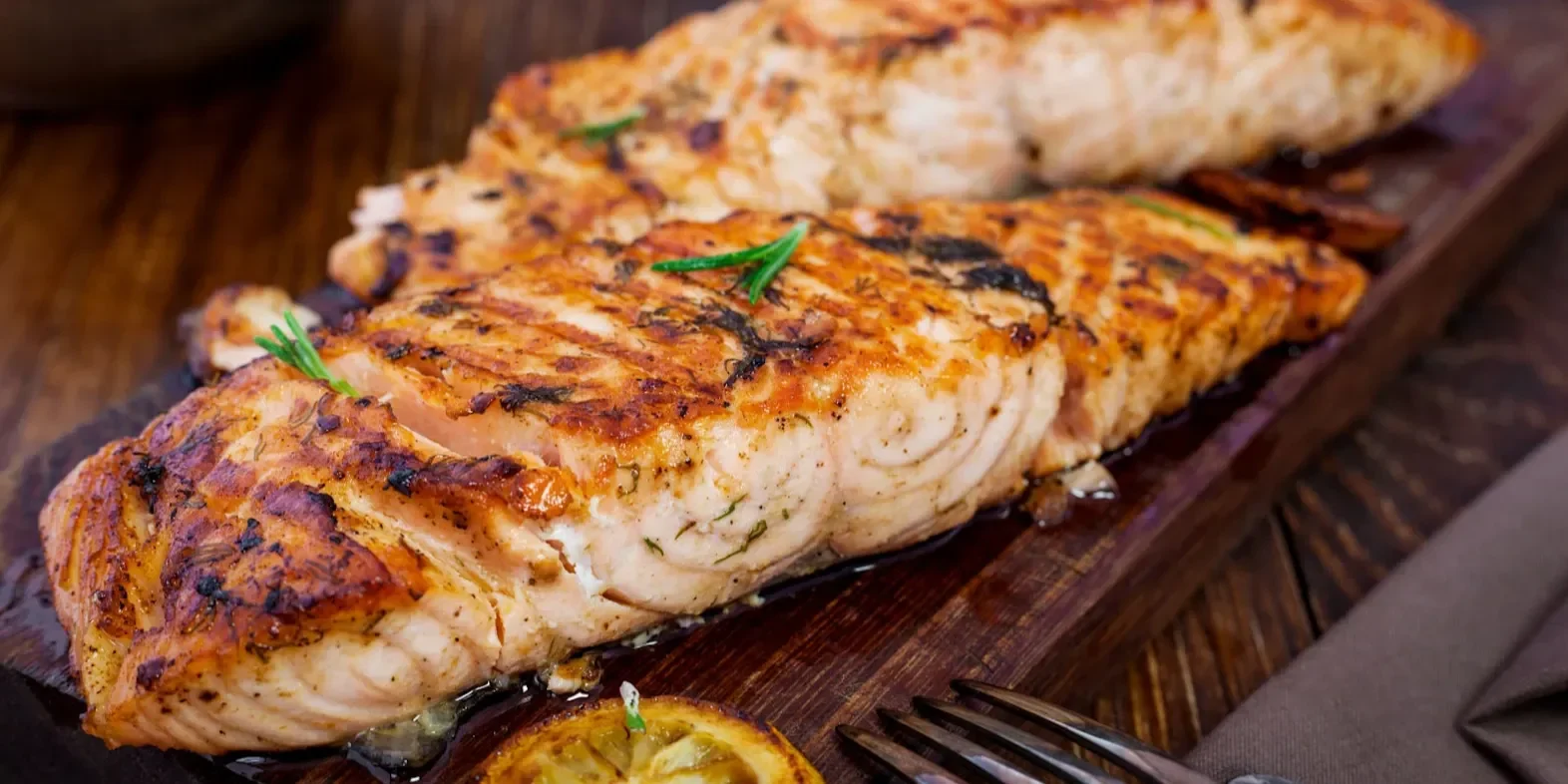 Grilled salmon fillets on a wooden board with herbs and charred lemon slices.