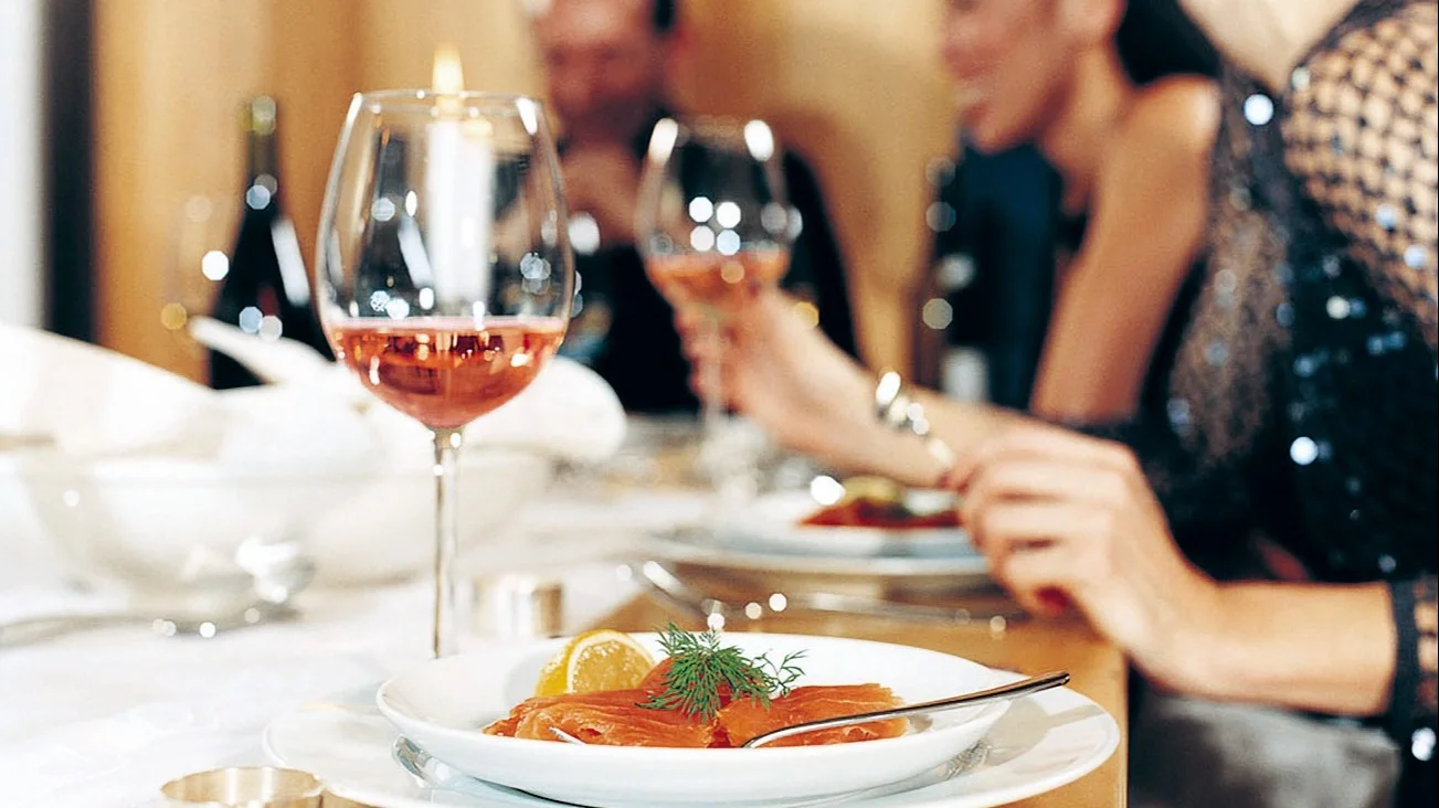 A glass of rose on a fancy dining table paired with a white platter of salmon garnished with a slice of lemon and herbs.