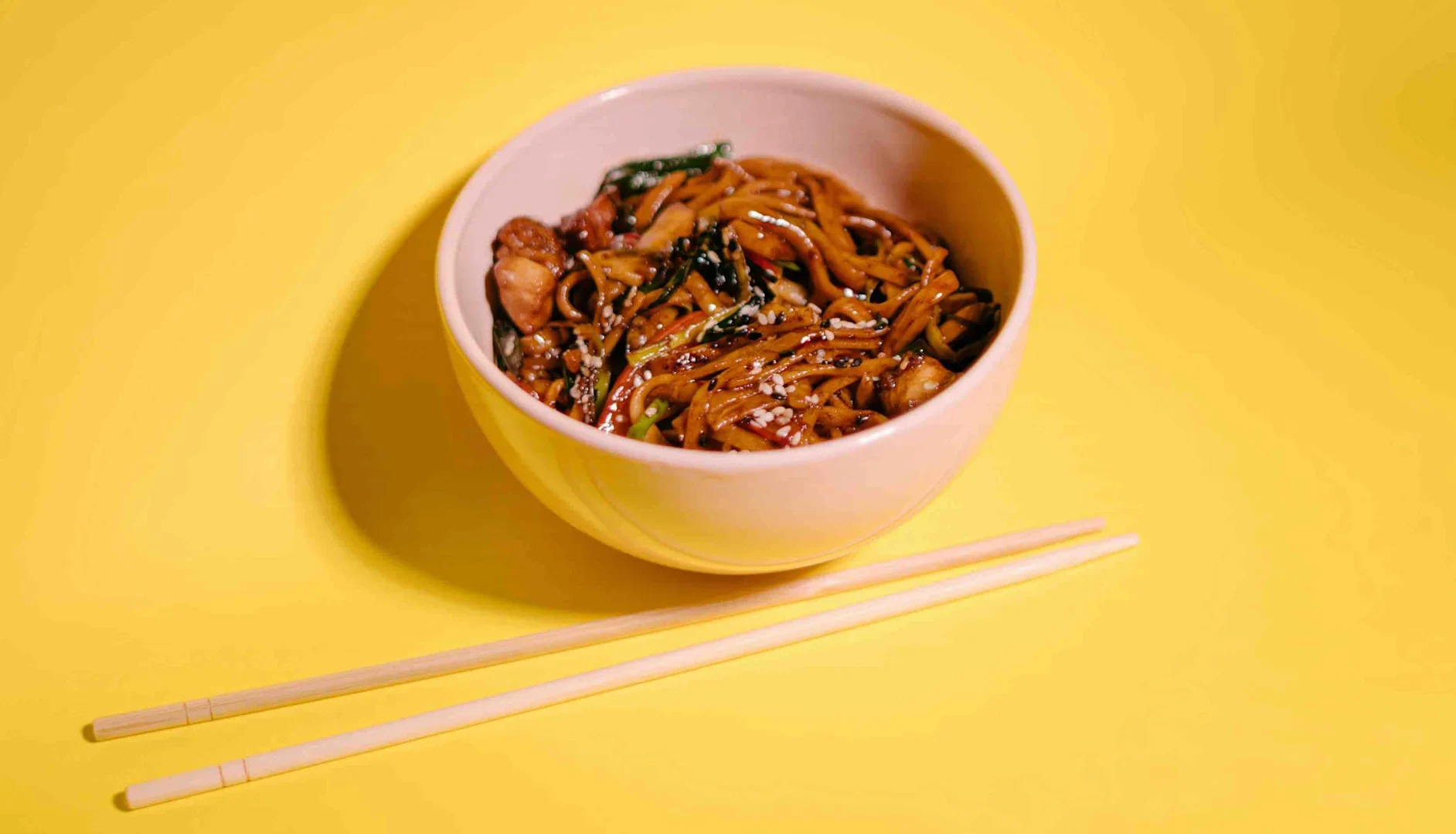 Bowl of stir-fried noodles with vegetables and sesame seeds on a yellow background with chopsticks.