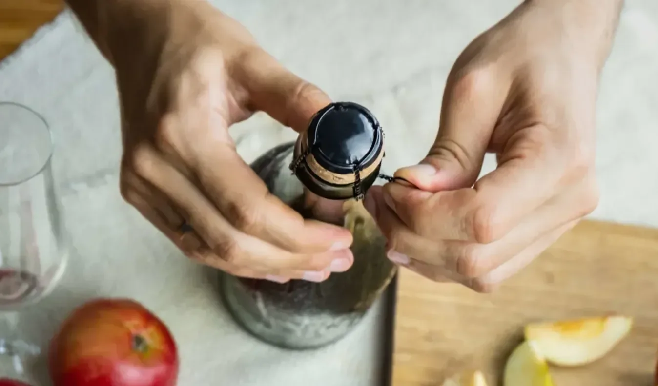 Hands opening the wire cage on a sparkling wine bottle, with apples, a glass, and sliced fruit arranged on a wooden board nearby.