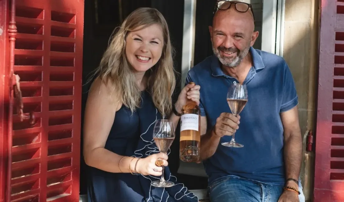A man and a woman stood in-front of a red shuttered window holding a bottle of rose wine, smiling happily at the camera.
