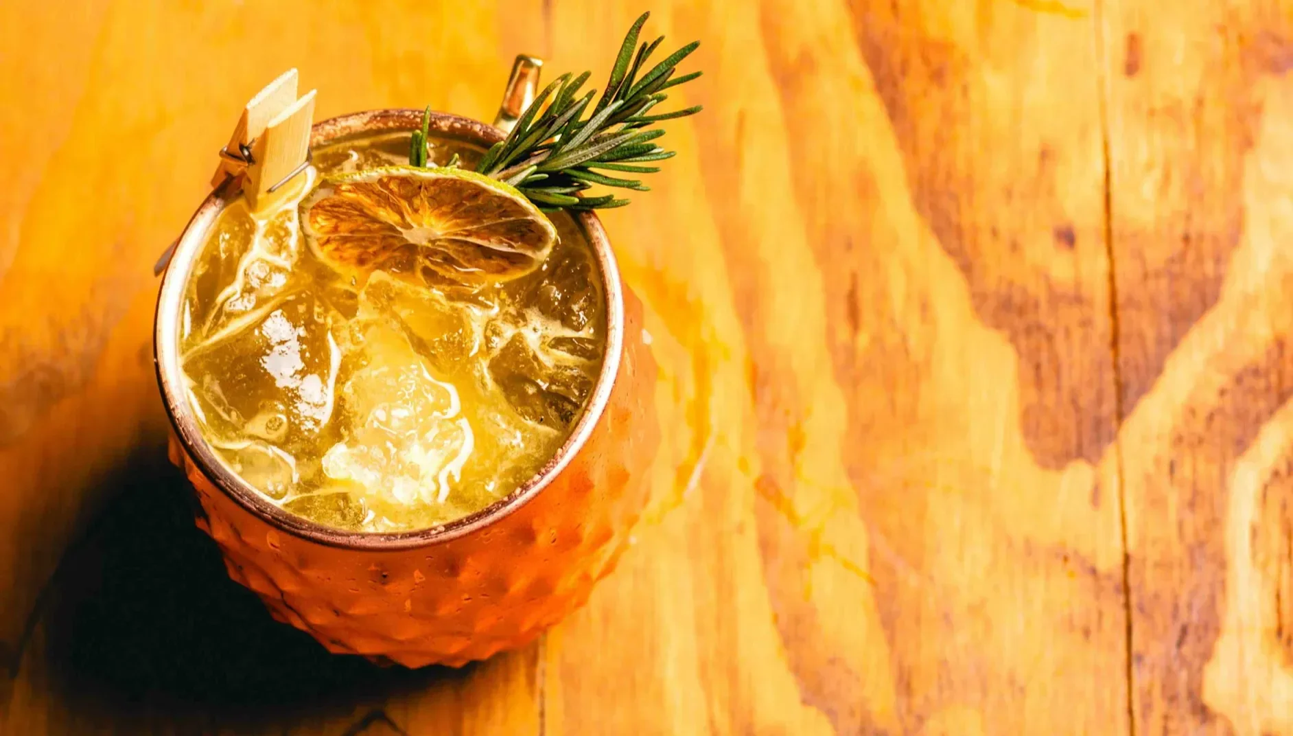 Iced cocktail in a textured copper mug topped with dried citrus and a sprig of rosemary on a wooden surface.