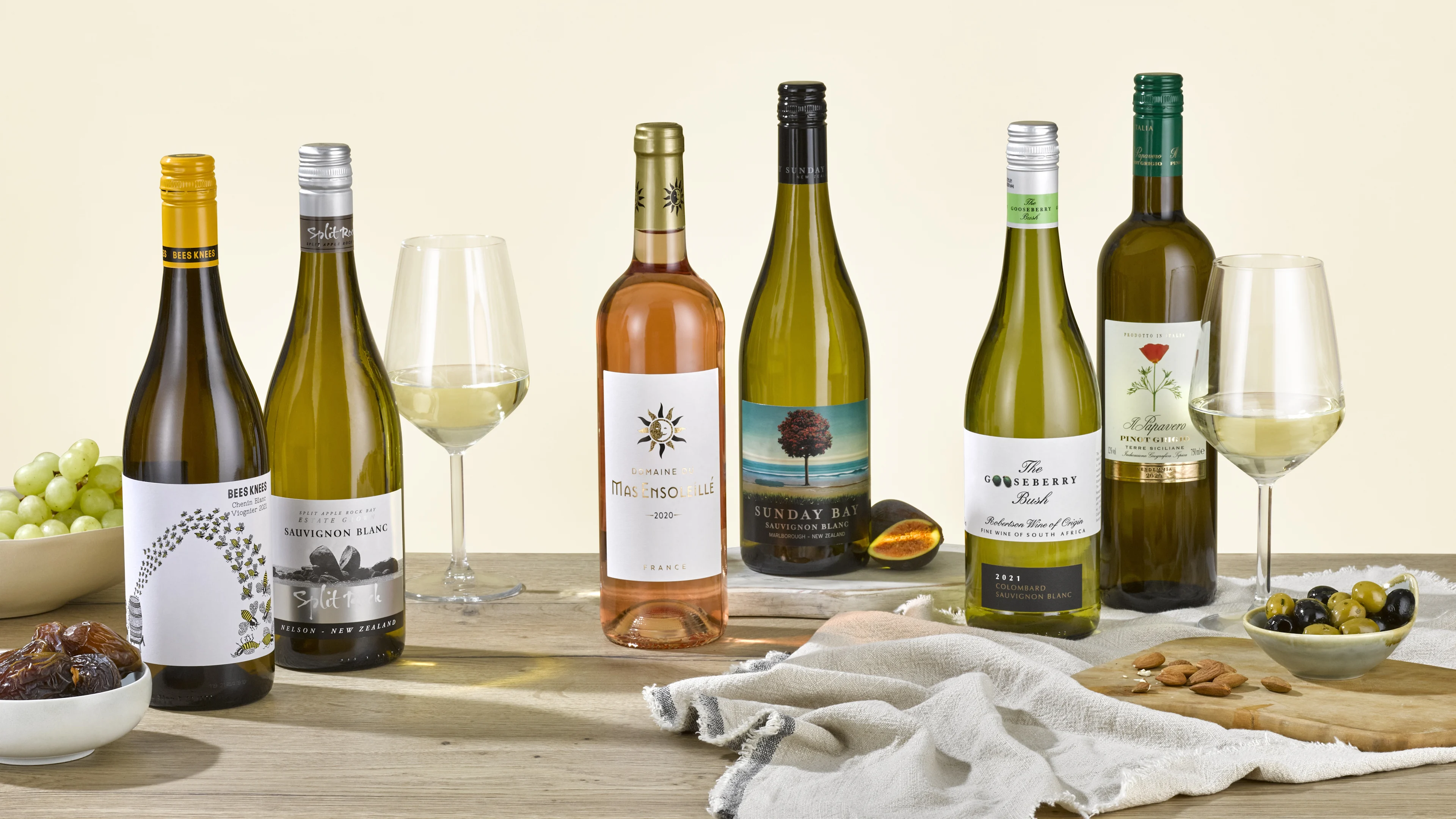 Selection of white wine bottles with filled wine glasses, grapes, olives, dates and almonds on a wooden table.