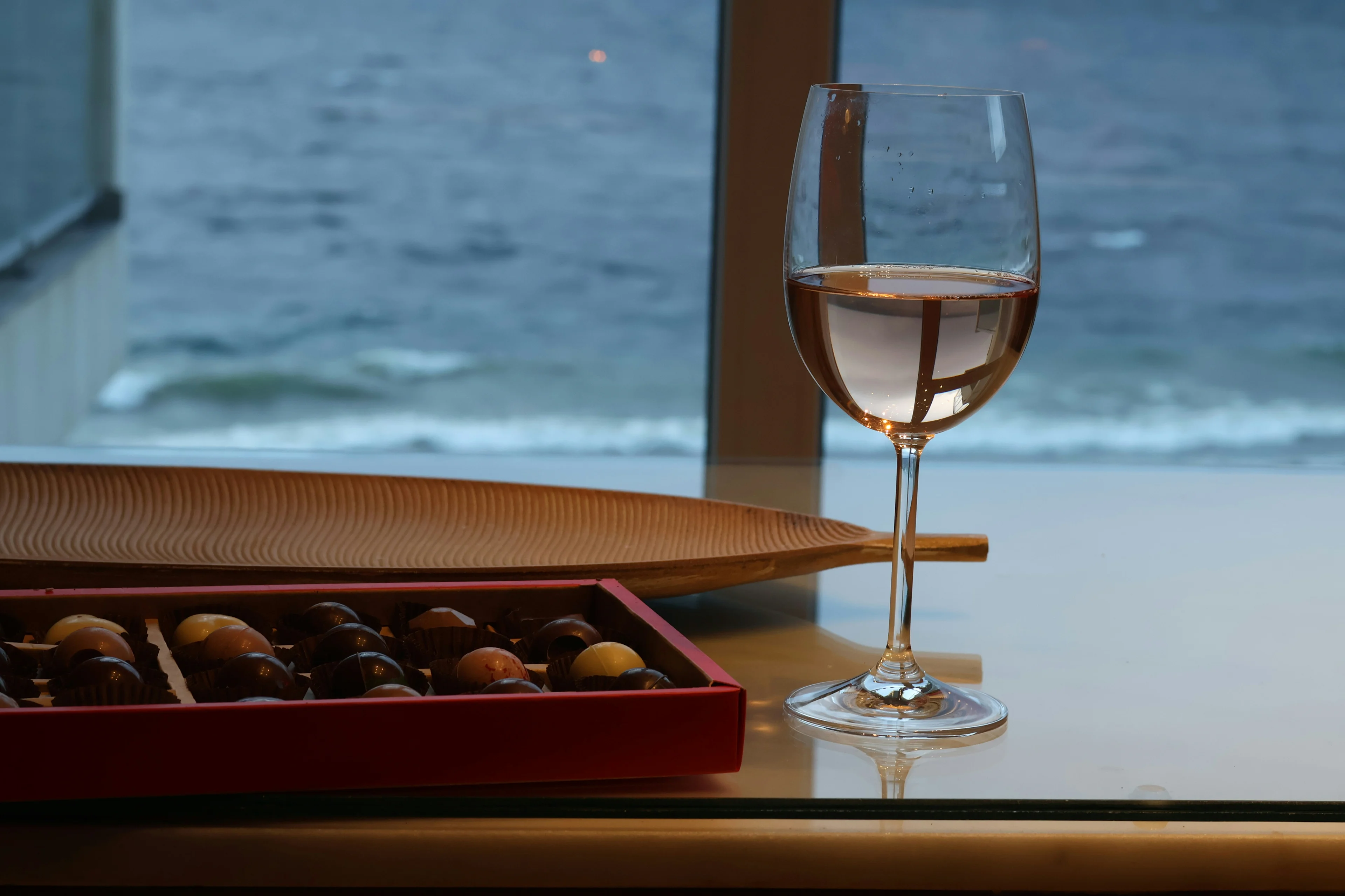 Glass of rosé wine on a table beside a box of chocolates with the ocean visible through the window.