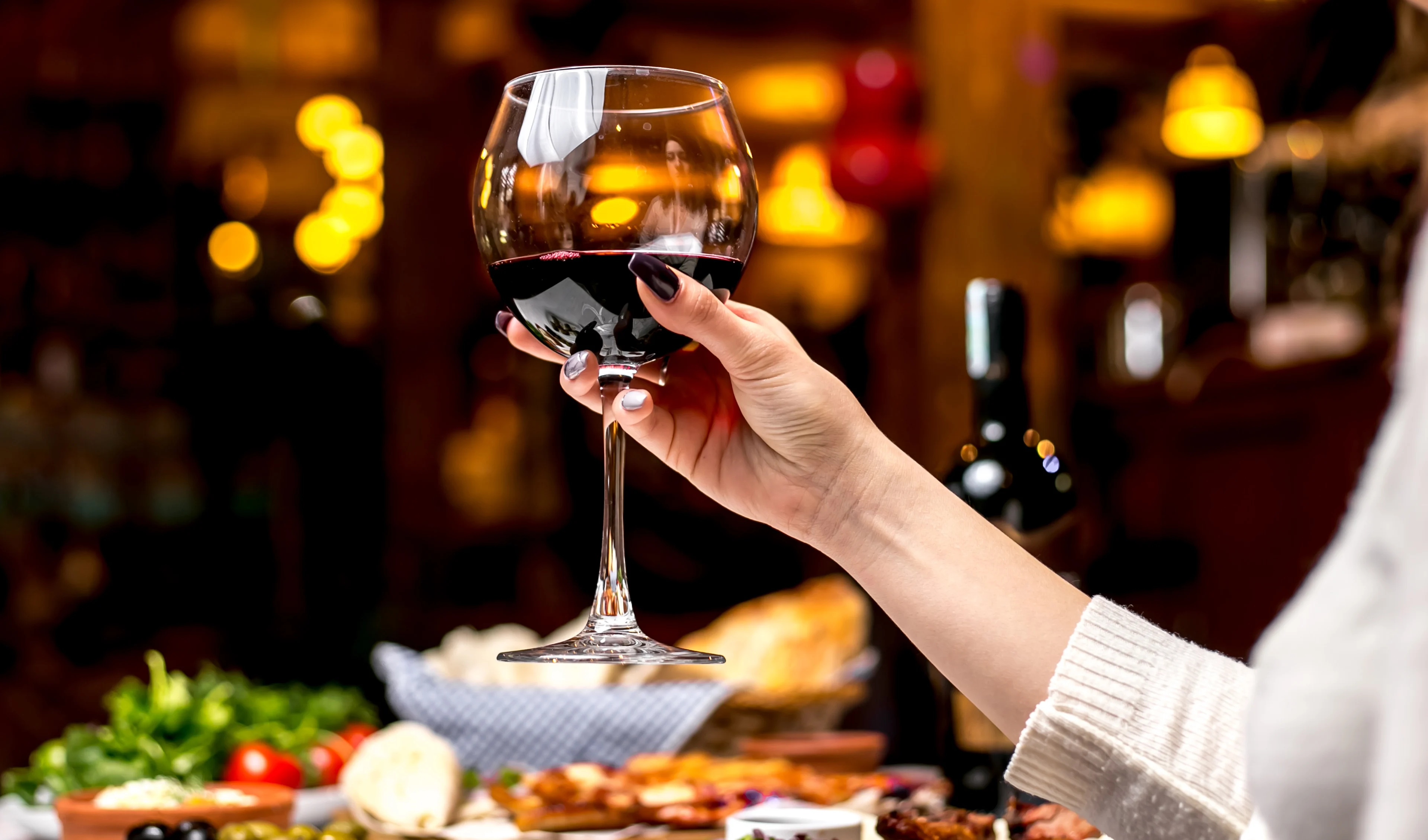 Close-up side view of a woman's arm at a dinner table holding a large glass of fruity red wine, with a blurred background of vegetables, salad and wine bottle on the table.