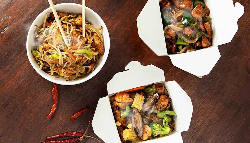 Bowls and takeout boxes of stir-fried noodles and vegetables with chopsticks on a wooden table.