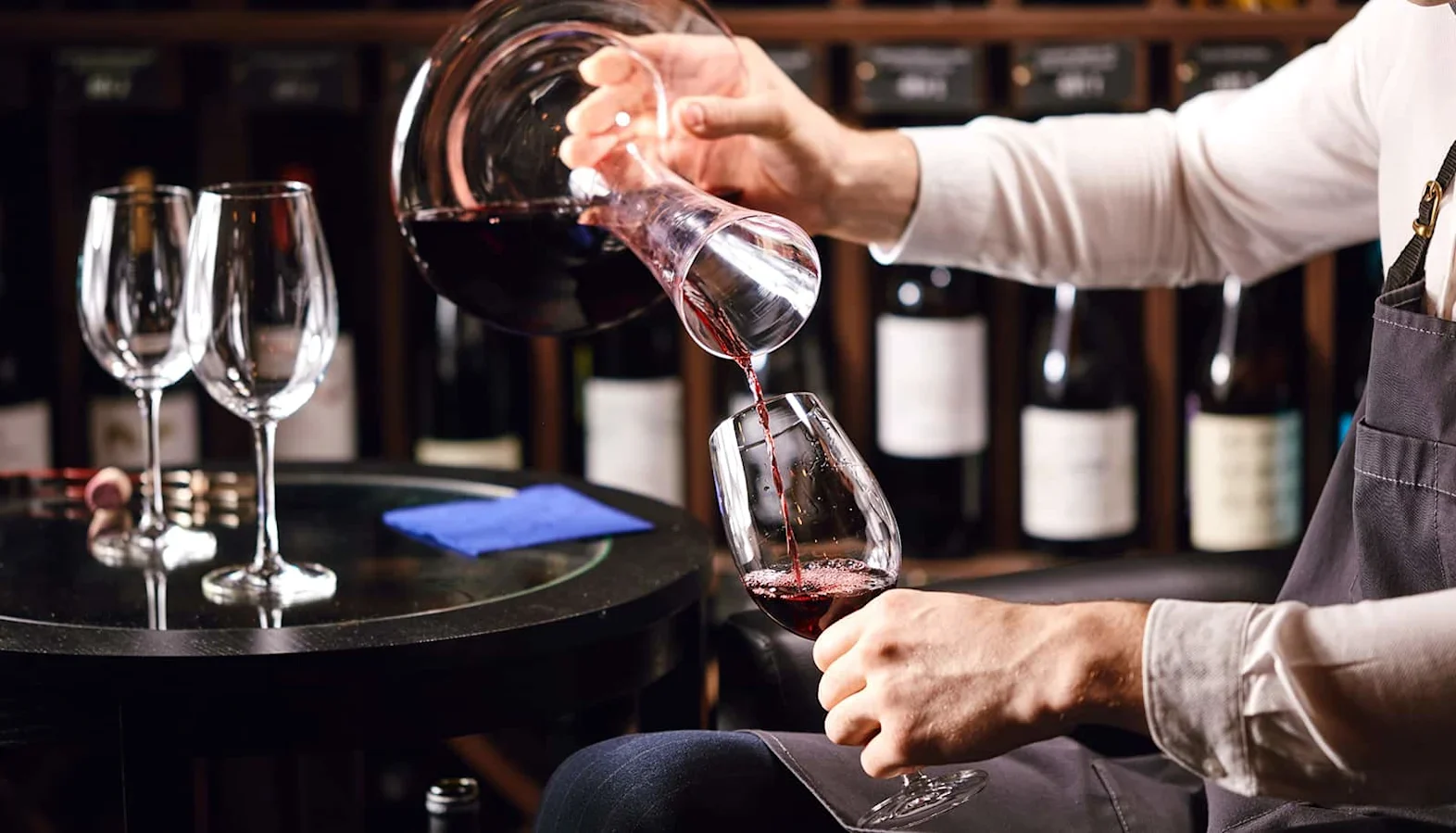 Person pouring red wine from a glass decanter into a wine glass on a table with empty glasses and wine bottles in the background.