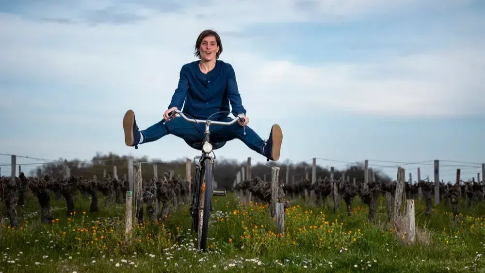 Person riding a bicycle between vineyard rows with legs outstretched above a flower-filled grass path.