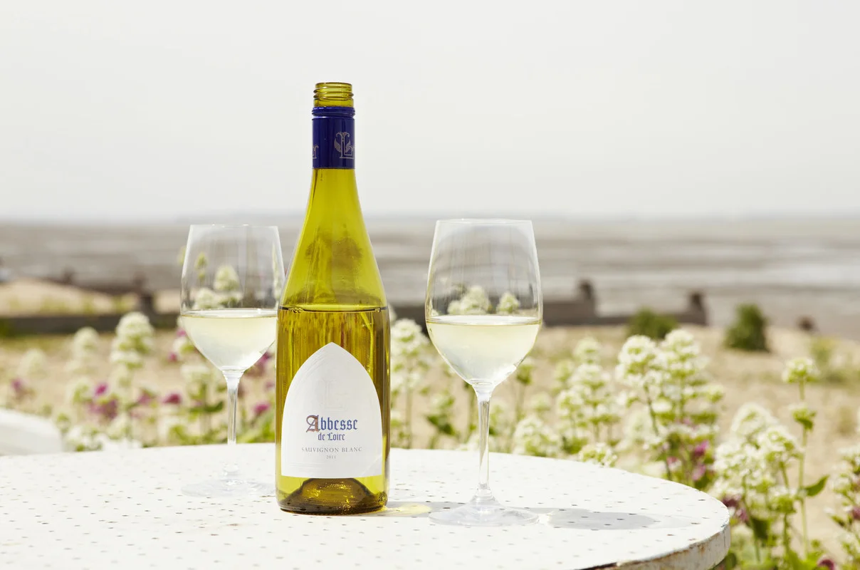 Bottle of Abbesse de Loire Sauvignon Blanc with two glasses of white wine on a white table overlooking the coastal landscape of Kent.