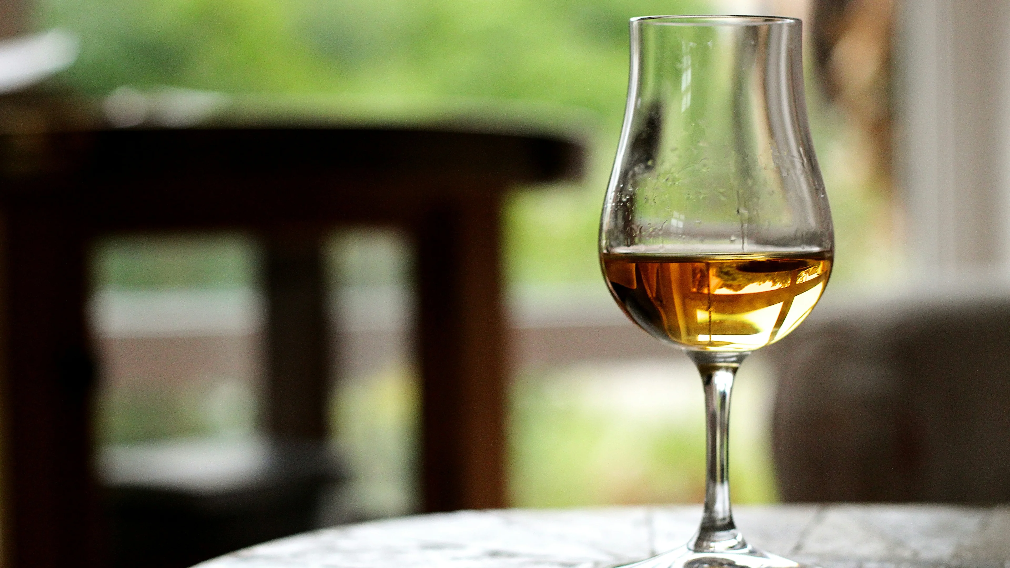 Glass of amber-coloured wine on a round table indoors with softly blurred greenery outside.