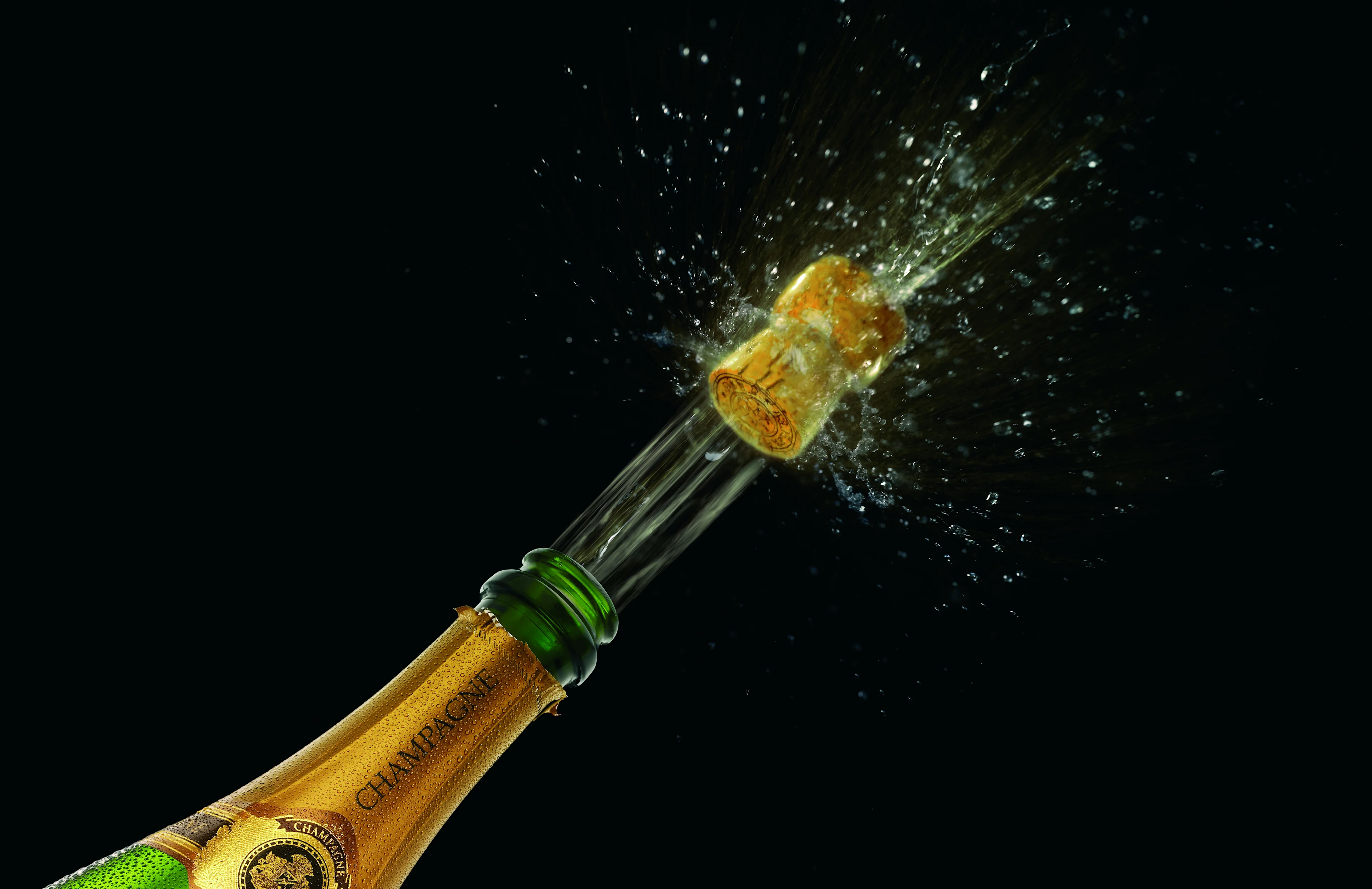 A green bottle of champagne cropped to the top top shop the golden label at the top of the bottle, and the cork popping off, with small splashes of champagne exploding from the bottle.
