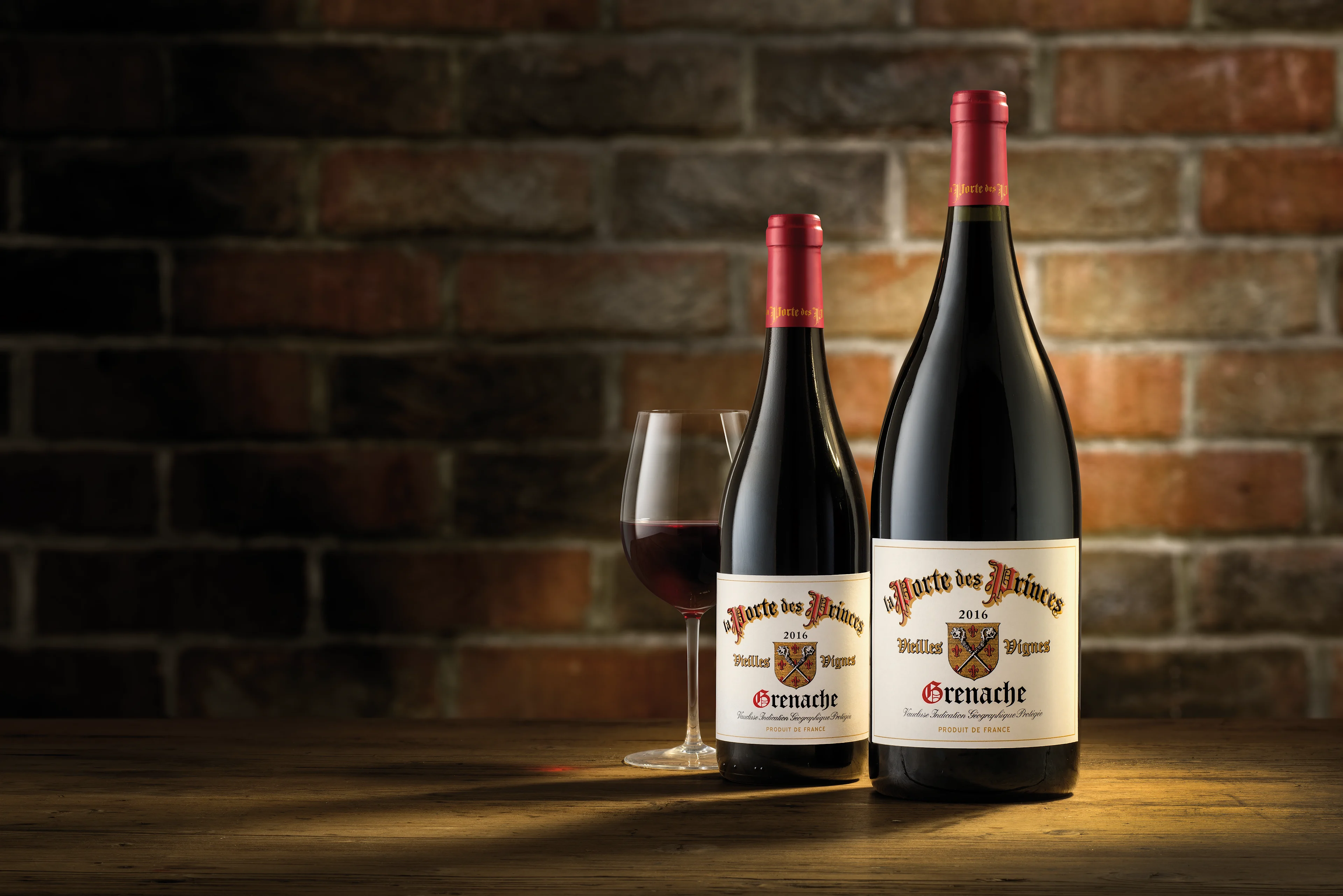 Two bottles of red wine with a filled wine glass placed on a wooden table against a brick backdrop.